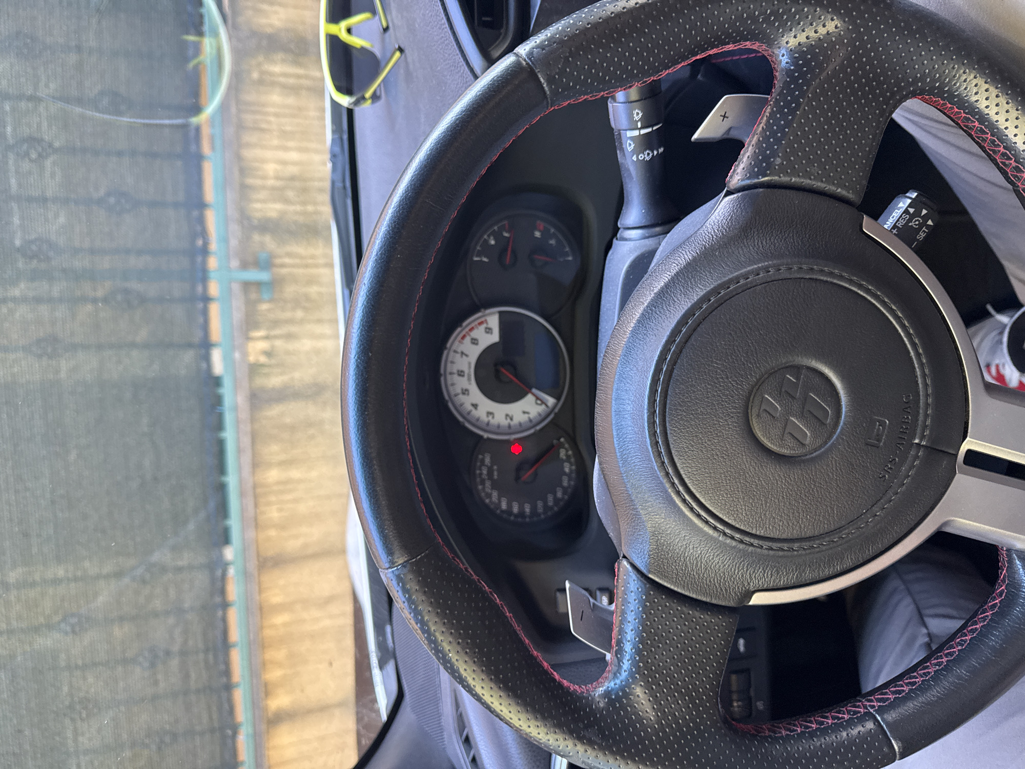 The image shows the interior of a car, focusing on the steering wheel and dashboard. The steering wheel is black with red stitching and has paddle shifters. The dashboard includes a speedometer and tachometer. A pair of neon yellow sunglasses is resting on the dashboard. Outside the car, a black mesh fence is visible above a concrete wall. The scene suggests the car is parked outdoors in a location with a fence and wall. The sunglasses indicate a sunny environment. There are no people clearly visible in the image, but the perspective suggests the photograph was taken from the driver's seat. The overall impression is of a parked car, possibly on a warm day.