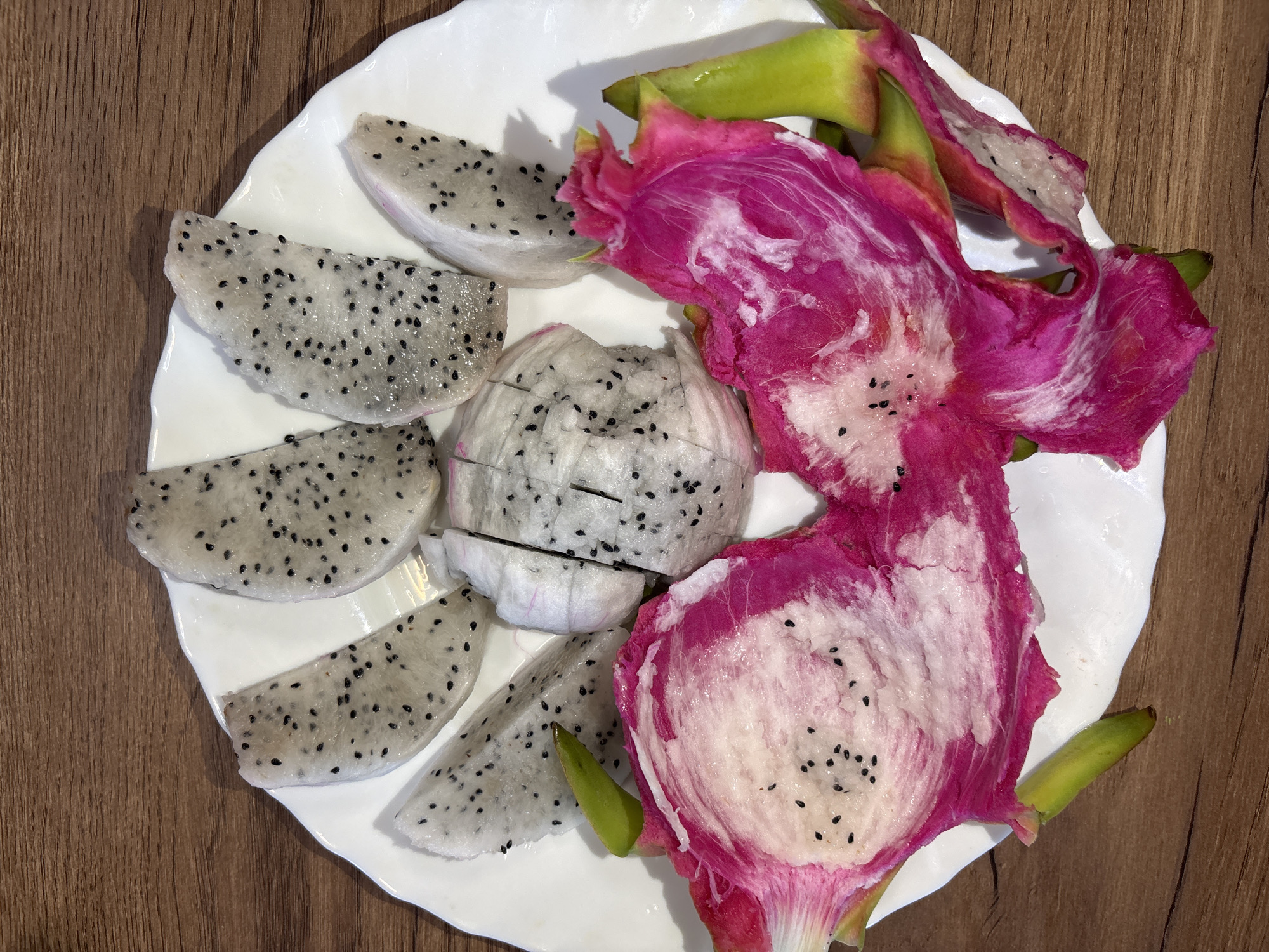 A white plate of dragon fruit is on a wood table. Slices of dragon fruit are on the left side of the plate, and the skin of the dragon fruit is on the right side of the plate. There are no people in the image. The photo was taken inside. The exact location is unknown. The location is not identified as Izhevsk, Russia.