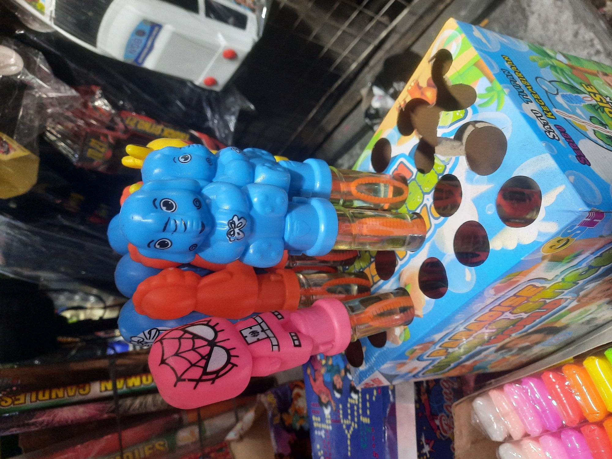 In North Jakarta, Indonesia, this image captures a close-up view of a display of bubble wands. The wands, some featuring character designs like Spider-Man and elephants, are inserted into a colorful cardboard box decorated with cartoon imagery and text that reads "Satu Tiupan Seribu Kegembiraan" and "PARADISE". The wands have clear plastic tubes containing orange liquid, presumably the bubble solution. In the background, various items are visible, including what appear to be fireworks or sparklers, other toys, and packaging, all suggesting a retail or market setting. The lighting is somewhat dim, with a focus on the foreground objects.