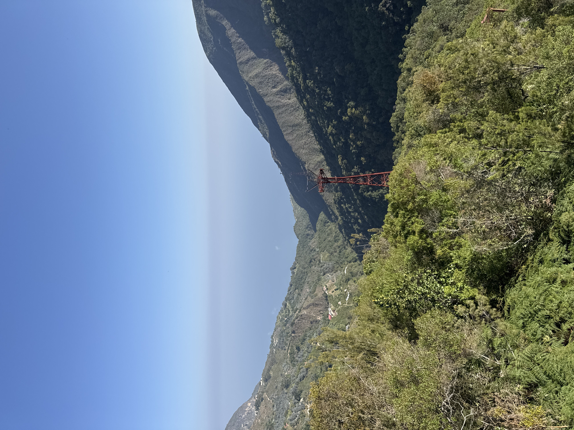The image shows a view in the city of San Antonio de Galipán, Venezuela. In the background, there is a tall mountain and a deep ravine with a red metal structure (likely a part of a tram or cable car system) spanning across it. The foreground is filled with dense green vegetation, including trees and shrubs. The sky is a clear blue with a slight haze near the horizon. The photo is taken during daylight, evidenced by the lighting. There are no people visible in the image.