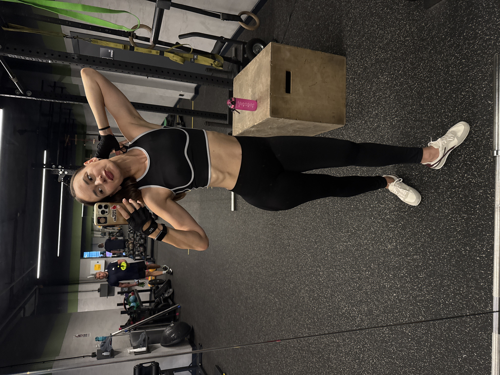 In a gym environment, a woman is taking a selfie in front of a mirror. She is wearing a black sports bra and black leggings, with white and grey athletic shoes. She also has on black fingerless gloves and a black fitness tracker on her left wrist. Her right hand is behind her head, and her left hand is holding her phone. A pink water bottle sits on a wooden plyometric box to her right. In the background, through the mirror's reflection, a man in a dark blue t-shirt and shorts is visible, holding a yellow object. The gym has a dark, speckled floor and industrial-style equipment, including weight racks and suspension trainers. The woman is positioned in the foreground, with the gym equipment and the man in the reflection forming the background. This scene is located in the city of Sochi, Russia.