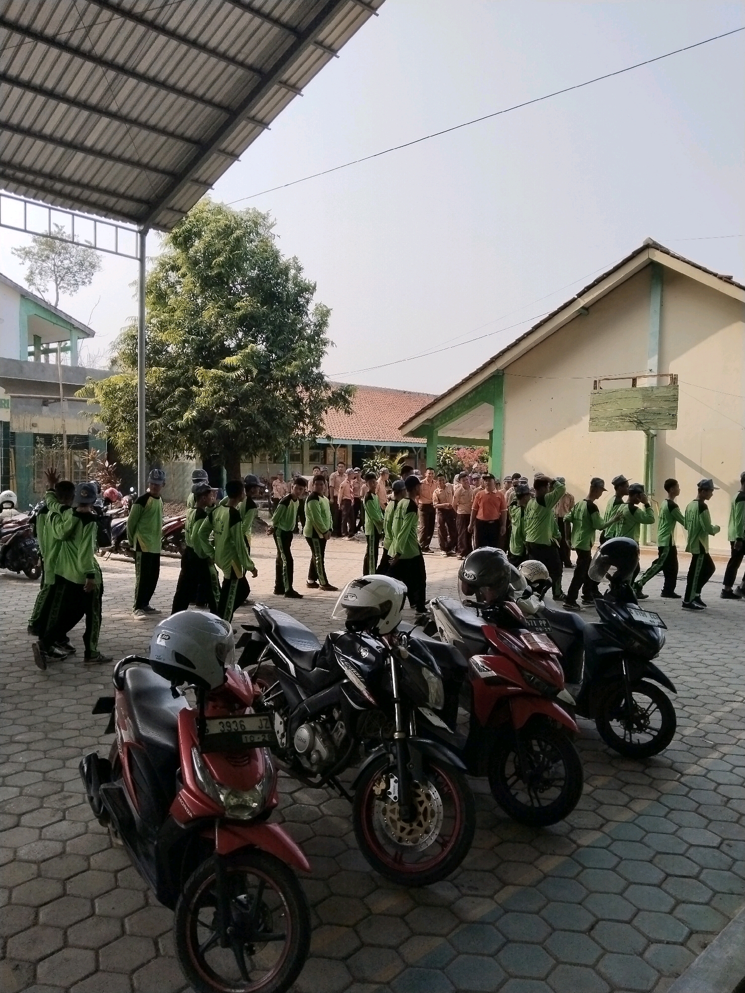 The image shows a group of students in green uniforms gathered in an outdoor courtyard, possibly at a school. Several motorcycles are parked in the foreground. The environment appears to be in Pakulaut, Indonesia, characterized by buildings with tiled roofs and a paved courtyard with a honeycomb pattern. Some students are interacting or observing, while others are standing still. The scene is lit by natural daylight, suggesting it is daytime.