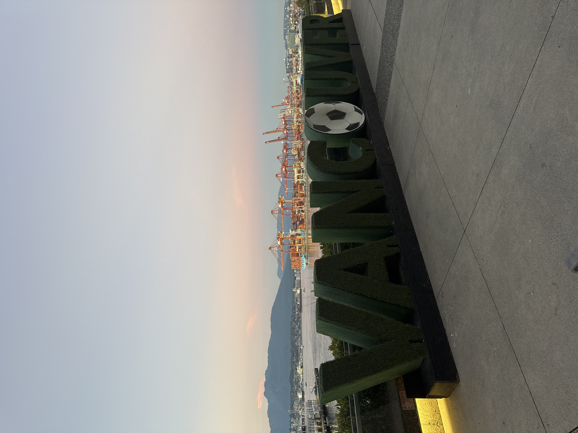 An exterior, eye-level, wide shot captures a large, green, artificial turf sign spelling out "VANCOUVER" with a soccer ball replacing the letter "O." The sign is situated in the foreground, facing the viewer, and is positioned on a dark base against a concrete walkway. Behind the sign, a panoramic view of Vancouver, Canada, unfolds.
In the middle ground, a body of water, likely the Vancouver Harbour, reflects the soft twilight sky. The water is dotted with various vessels, including cargo ships and smaller boats. Dominating the skyline are numerous red cranes at a port, stacked with shipping containers, suggesting a bustling industrial area. Beyond the port, a distant city skyline is visible, with buildings stretching across the horizon. In the far distance, a mountain range forms a majestic backdrop, its peaks softened by the fading light.
The sky above is a gradient of pale blue at the top, transitioning to hues of pink and orange near the horizon, indicative of either sunrise or sunset. The lighting on the sign is provided by subtle yellow accent lights along its base, casting a warm glow on the concrete. The overall atmosphere is serene and picturesque, showcasing a blend of urban infrastructure, natural beauty, and a prominent city landmark. There are no people visible in the frame.