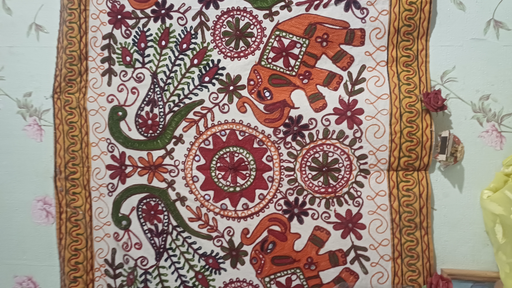 This image shows a close-up view of an embroidered textile, likely a wall hanging or tapestry. The textile features a repeating pattern of stylized motifs, including elephants and peacocks, rendered in various shades of orange, green, red, and brown embroidery on a cream-colored background. Intricate floral designs and circular medallions are also prominent. The textile is bordered by a decorative trim. In the background, a pale green wall with a floral wallpaper pattern is visible, along with some decorative fabric elements that appear to be artificial flowers. There is no indication of the city of Izhevsk, Russia, or any specific location within it. The image is static and does not depict any actions or interactions.