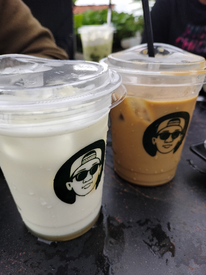 This close-up shot captures two iced beverages in clear plastic cups placed on a dark, possibly metallic or stone, table in an outdoor setting in Dramaga, Indonesia. The cup on the left contains a light-colored, possibly milky, drink with ice, while the cup on the right holds an iced coffee with a black straw protruding from its clear dome lid. Both main cups feature an identical black and white logo: a cartoonish illustration of a person's head wearing a backwards baseball cap and sunglasses. The table surface beneath the cups shows signs of condensation or moisture, hinting at cold drinks or a humid environment. In the softly blurred background, a person's brown-sleeved arm is visible alongside lush green foliage and another distant plastic cup with a green-tinged beverage, suggesting a cafe or casual eatery. The natural, bright lighting indicates it is daytime. No discernible text is present, apart from the distinctive visual logo on the cups.