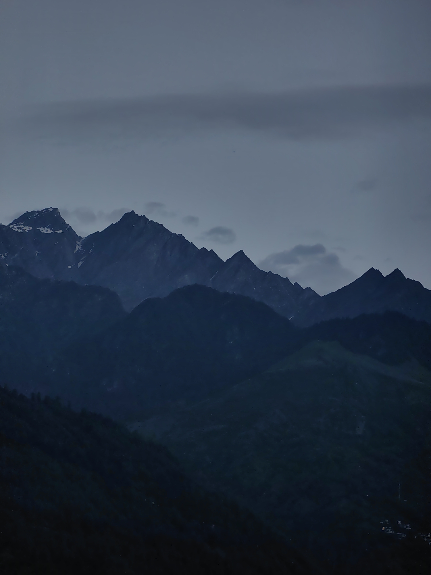 The image is a dark, moody landscape photograph featuring a series of silhouetted mountain ranges. The sky is overcast with heavy, dark clouds, creating a somber atmosphere. The mountains themselves are deep blue-black, with the peaks in the background appearing fainter and more diffused due to atmospheric perspective. Some of the higher peaks in the foreground show hints of snow or lighter rock, particularly on the left side. The foreground consists of darker, forested mountain slopes that fade into near blackness at the bottom of the frame. There is a faint suggestion of human settlement in the very bottom right corner, with a few small structures visible. The overall impression is one of a dramatic and somewhat desolate mountain scene, likely taken at dusk or during heavy cloud cover.
The image does not contain any discernible elements that would allow for a precise location within Vashisht, India. Without further contextual information or distinctive landmarks, it is impossible to definitively pinpoint the location within Vashisht based solely on this photograph.