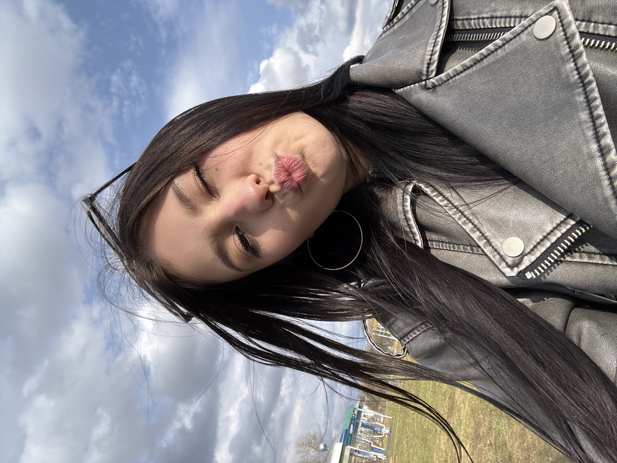 A young woman with long, dark hair is posing for a selfie outdoors. She is wearing a gray leather jacket and a large hoop earring. Her eyes are partially closed, and she is puckering her lips. The background shows a partly cloudy sky and a grassy area with some buildings and structures that appears to be Фоки, Russia.