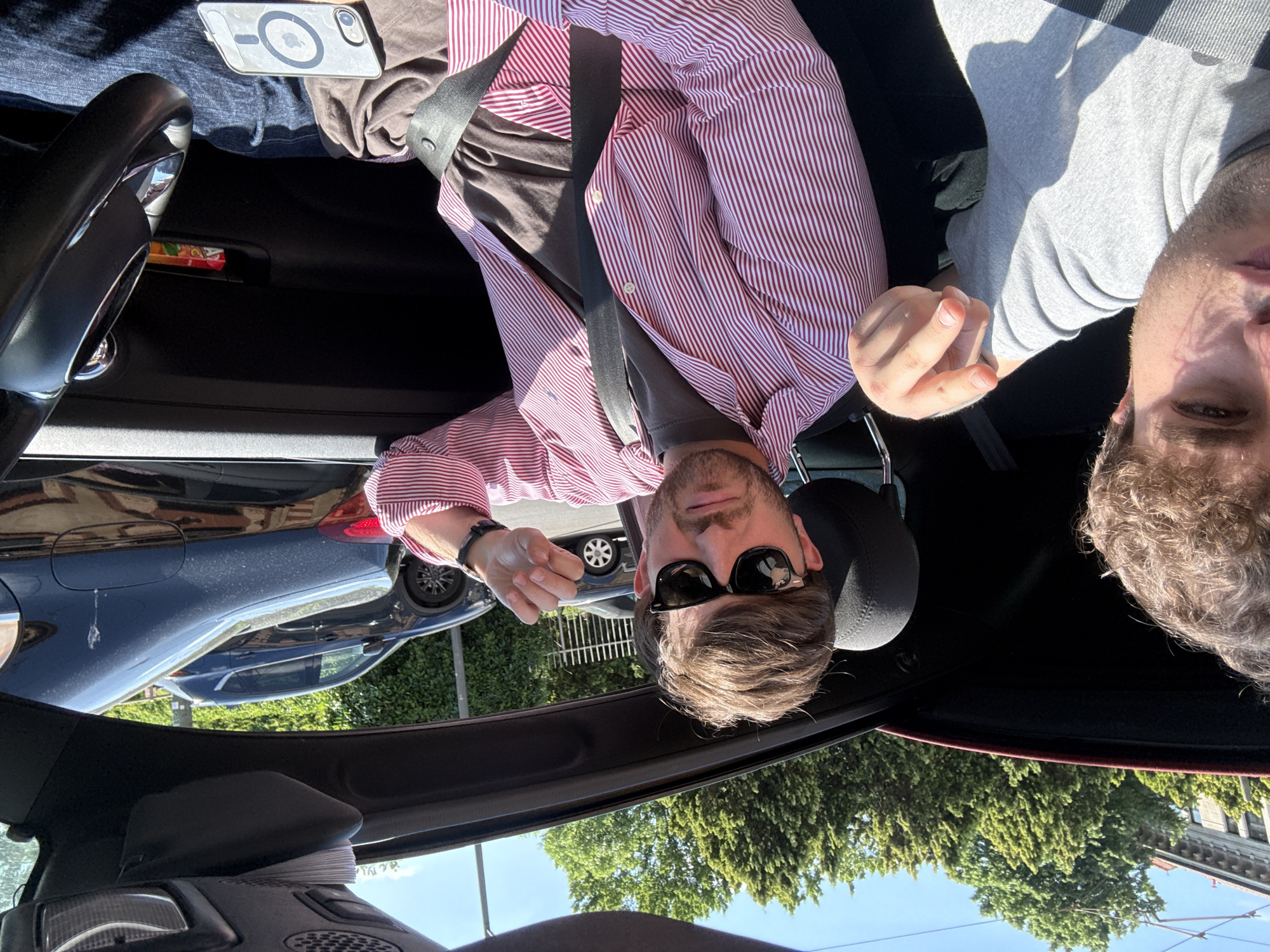 The photograph depicts two individuals inside a car. The person on the left is wearing a pink and white striped shirt, a dark t-shirt, and sunglasses, and is fastened in the car with a seatbelt. The other individual on the right is wearing a gray t-shirt. The car appears to be parked on a street with buildings and trees visible outside. Based on visual cues and the presence of European vehicles and architecture, the location is estimated to be in the city of Milan, Italy.