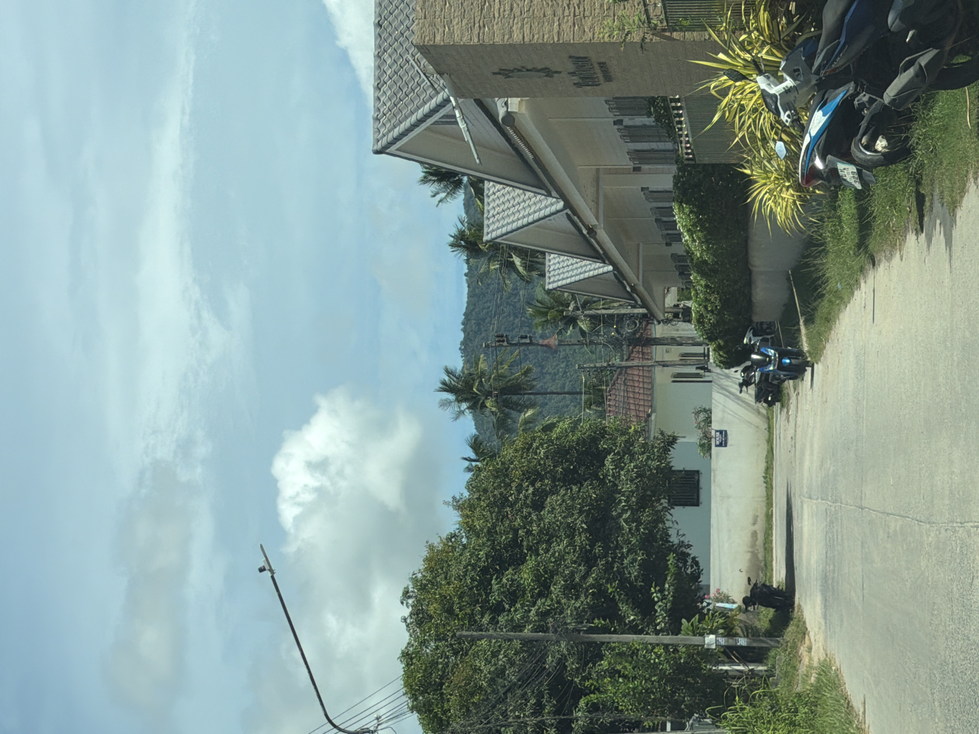 This media file depicts an outdoor scene of a street lined with buildings and lush vegetation. The perspective suggests the viewer is in a vehicle, looking out at the scenery.
The primary subjects are a series of modern buildings with tiled roofs and stone facades, suggesting a residential or commercial complex. In front of the buildings, there is a paved road with at least two motorcycles parked along the side. One motorcycle, closer to the foreground, is blue and black, while another, further down the road, is dark in color. Dense green trees and other plants flank the road and the buildings, with some tropical palm trees visible further in the background, suggesting a warm climate. A distant, verdant mountain range forms the backdrop. The sky is bright blue with scattered white clouds, indicating it is daytime with fair weather.
There is no clear activity or event taking place beyond the presence of parked vehicles. The scene conveys a sense of quiet or perhaps a pause in activity.
Notable details include the distinctive triangular roofing structure of the buildings and the sunny, clear weather. The presence of palm trees and the type of vegetation hint at a tropical or subtropical location. No specific time of day is indicated beyond it being daylight.
There is some visible text on one of the buildings. On the beige stone facade of the building on the right, there is a dark logo or sign with embossed lettering that appears to read "RIVA". Below this, there is smaller text, also embossed, that is difficult to read clearly but seems to start with "RESIDENCE". On a white wall of a building further down the street, there is a dark rectangular sign with white text that is also unreadable. The license plate of the blue and black motorcycle has Thai characters.