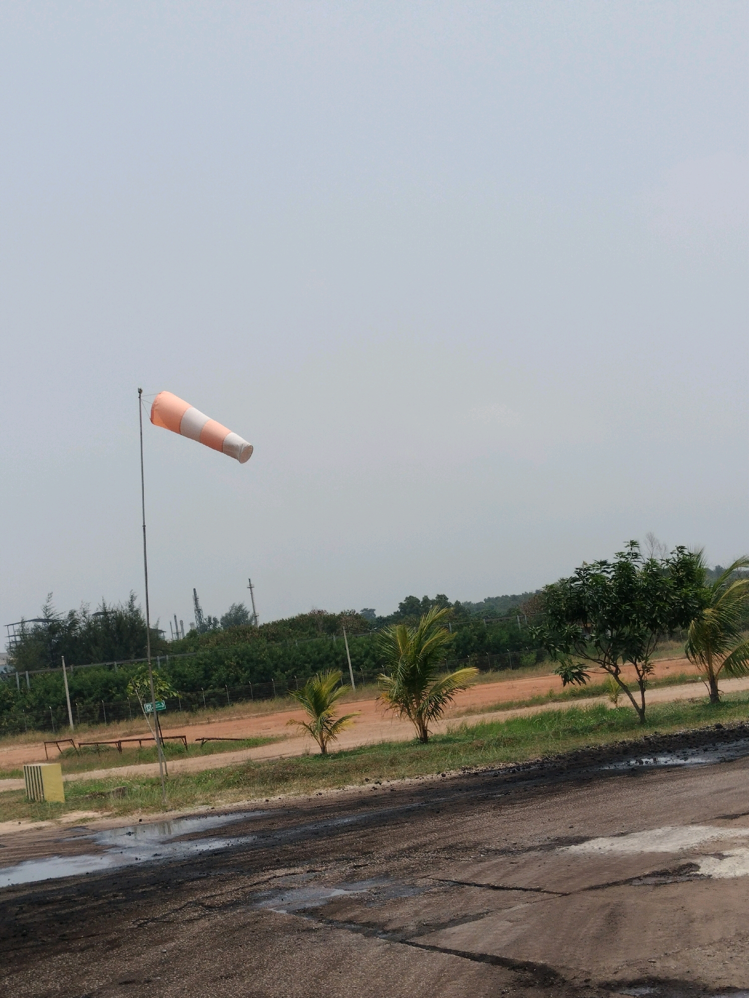 The image shows an orange and white windsock on a pole, indicating wind direction. It is set against a pale sky with a slightly hazy horizon. In the background, there is a line of green trees and some industrial structures, possibly including smokestacks. The foreground depicts a rough, dirt road with some patchy puddles and dark, muddy areas. There are also a few small palm trees and other vegetation scattered around the scene, as well as some park benches and a yellow concrete barrier in the middle ground. The overall impression is of an outdoor industrial or airport area under a dull sky. This scene could be located in an industrial zone within Batam, Indonesia, given the presence of industrial structures and the tropical vegetation.