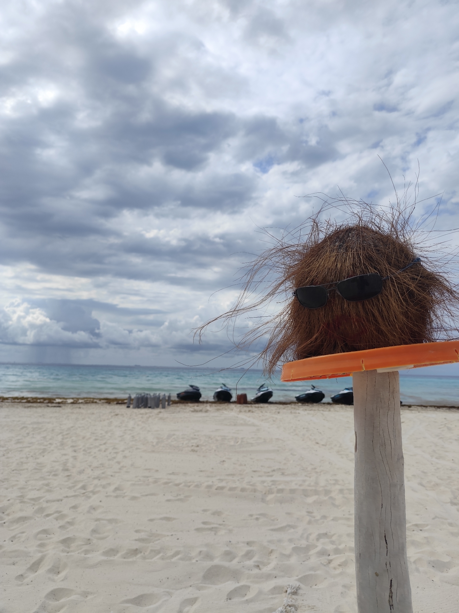 The scene is a beach in Playa del Carmen, Mexico. The foreground features a coconut with sunglasses, perched on an orange bar atop a white, weathered wooden post. The beach has light-colored sand with visible footprints. In the mid-ground, the ocean meets the beach with a line of seaweed. Behind that, several black jet skis are lined up along the shoreline. The sky is overcast with grey, stormy-looking clouds. Rain can be seen falling in the distance.