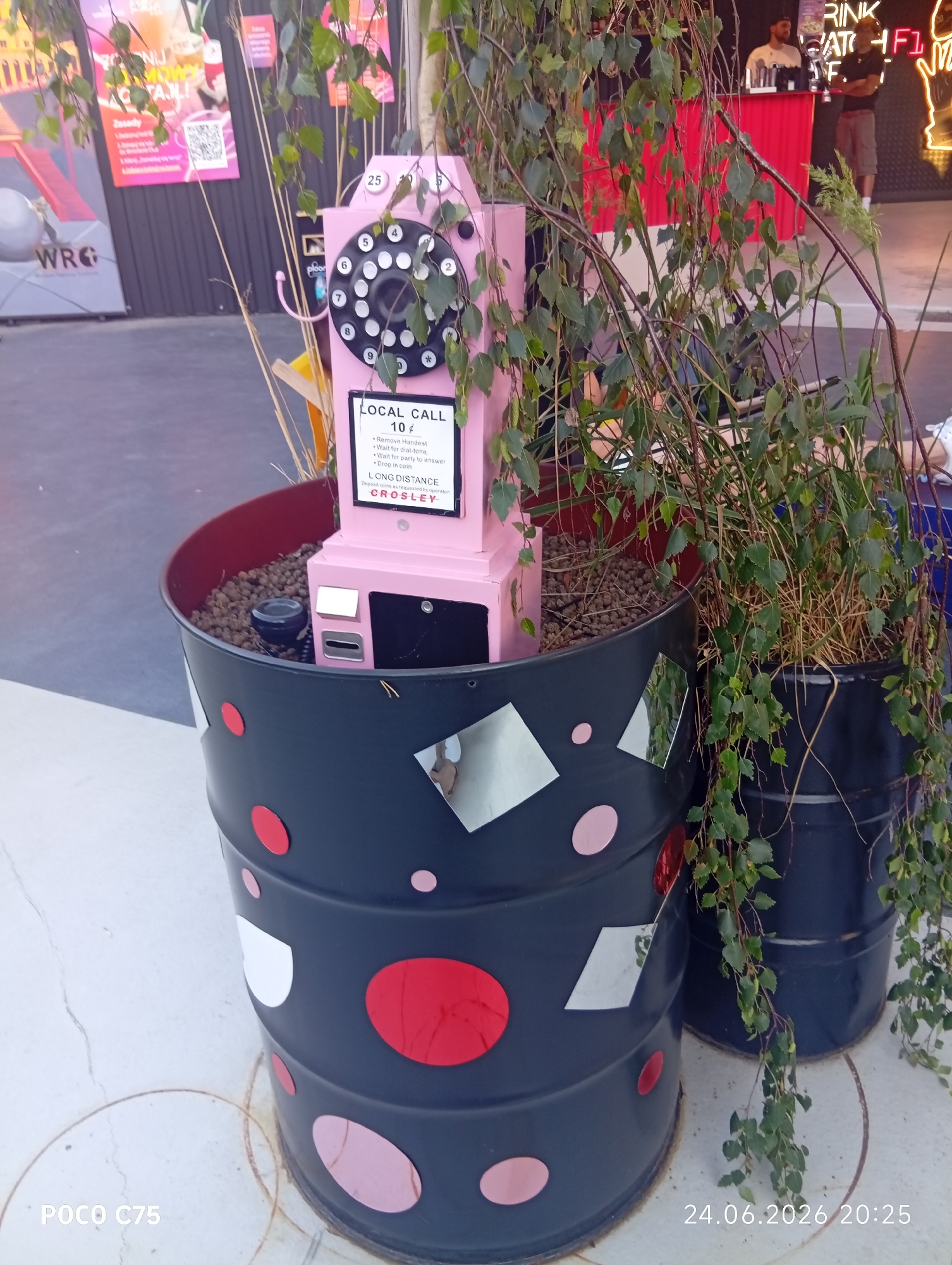 This media file features a close-up shot of a pink rotary dial telephone, repurposed as a decorative element, placed within a large black planter. The telephone is adorned with a sign that reads "LOCAL CALL 10¢" and provides instructions for its use, including "Remove Handset," "Wait for dial-tone," "Wait for party to answer," and "Drop in coin," along with information for "LONG DISTANCE." The planter itself is decorated with scattered circular and square reflective elements. Lush green foliage from a plant cascades over and around the telephone and planter, creating a natural, somewhat overgrown aesthetic.
In the background, hints of an outdoor venue are visible. A bar with a red counter is present, with two individuals standing behind it. To the left, a poster with the text "WRG" is partially visible. Neon lights and other signage suggest a vibrant, possibly entertainment-focused location. The overall scene appears to be outdoors, given the natural light and the presence of plants.
The timestamp at the bottom right corner indicates the image was taken on "24.06.2026 20:25," suggesting an evening setting. The visual elements, particularly the bar and signage in the background, imply a social gathering or a public space like a beer garden or an outdoor festival. No specific emotions are conveyed by the subjects, as the focus is primarily on the decorative telephone and planter. The location context of Wrocław, Poland, is provided separately.