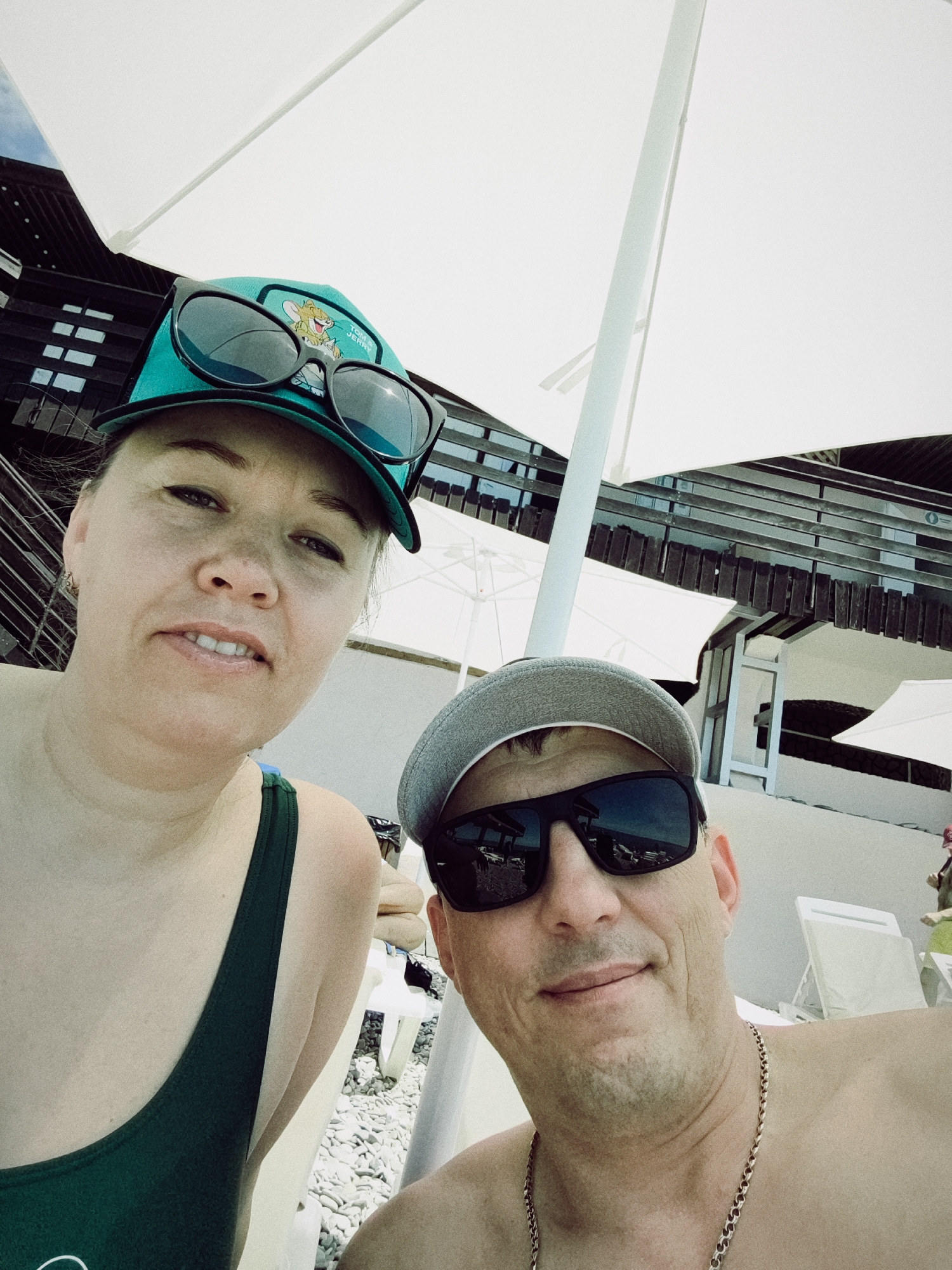 A selfie is taken from a low angle of a man and a woman. The man is in the foreground on the right, wearing a gray baseball cap, sunglasses, and a chain necklace. He is smiling. The woman is to his left, wearing a turquoise baseball cap with sunglasses perched on top, and a dark green swimsuit. She is also looking at the camera. Behind them, a white beach umbrella provides shade. In the background, there is a building with wooden slats and balconies, likely a hotel or resort. Beach chairs are visible on the pebbled ground. The overall setting suggests a vacation at a seaside location in Sochi, Russia.