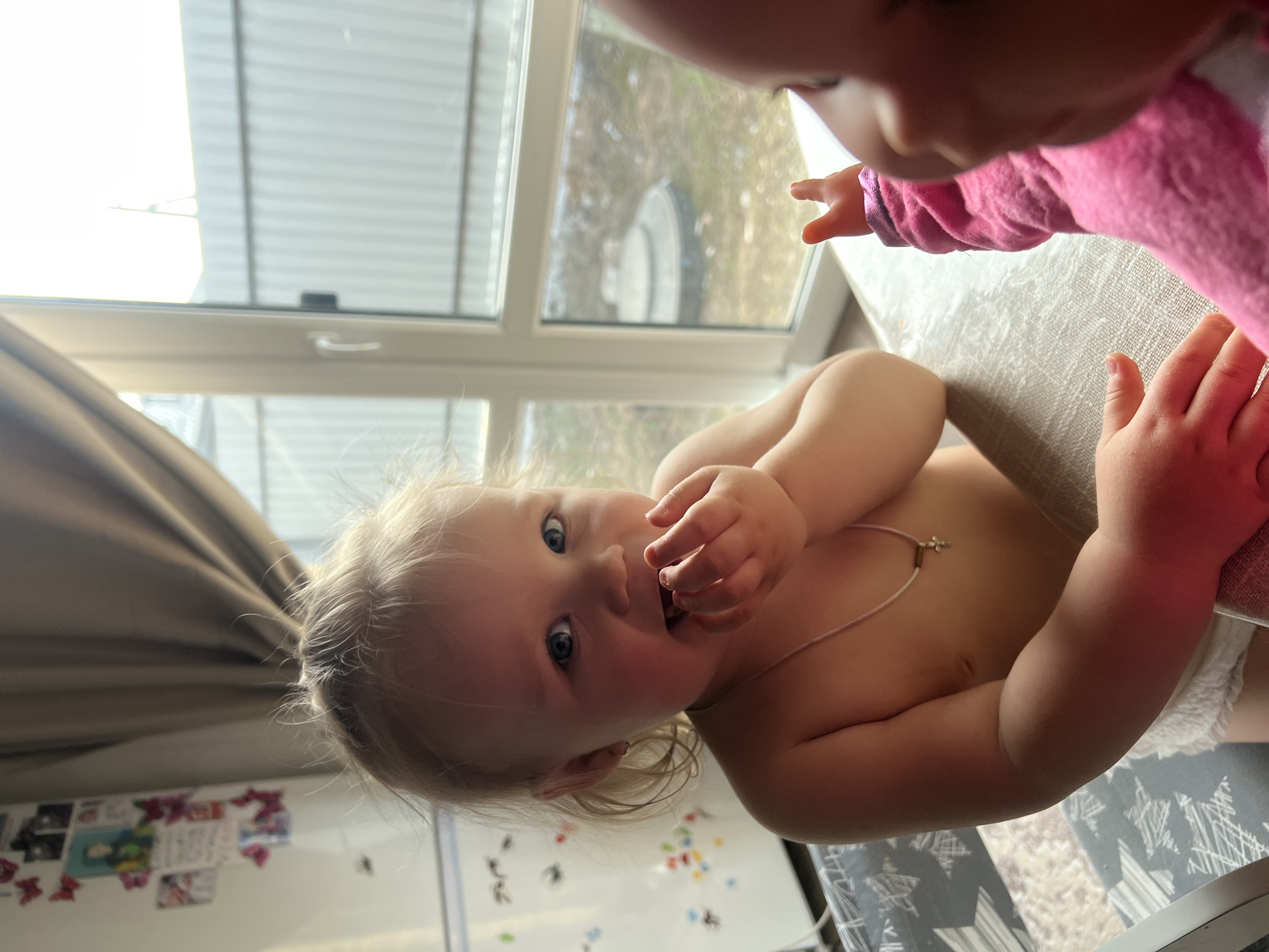 Two children interact indoors. One child, topless with blonde hair and blue eyes, looks at the camera while sitting at a table. They wear a thin necklace and a diaper. The other child, partially visible in a pink outfit, reaches a finger towards the blonde child. A window provides light in the background, and a decorated whiteboard can be seen in the bottom left corner.