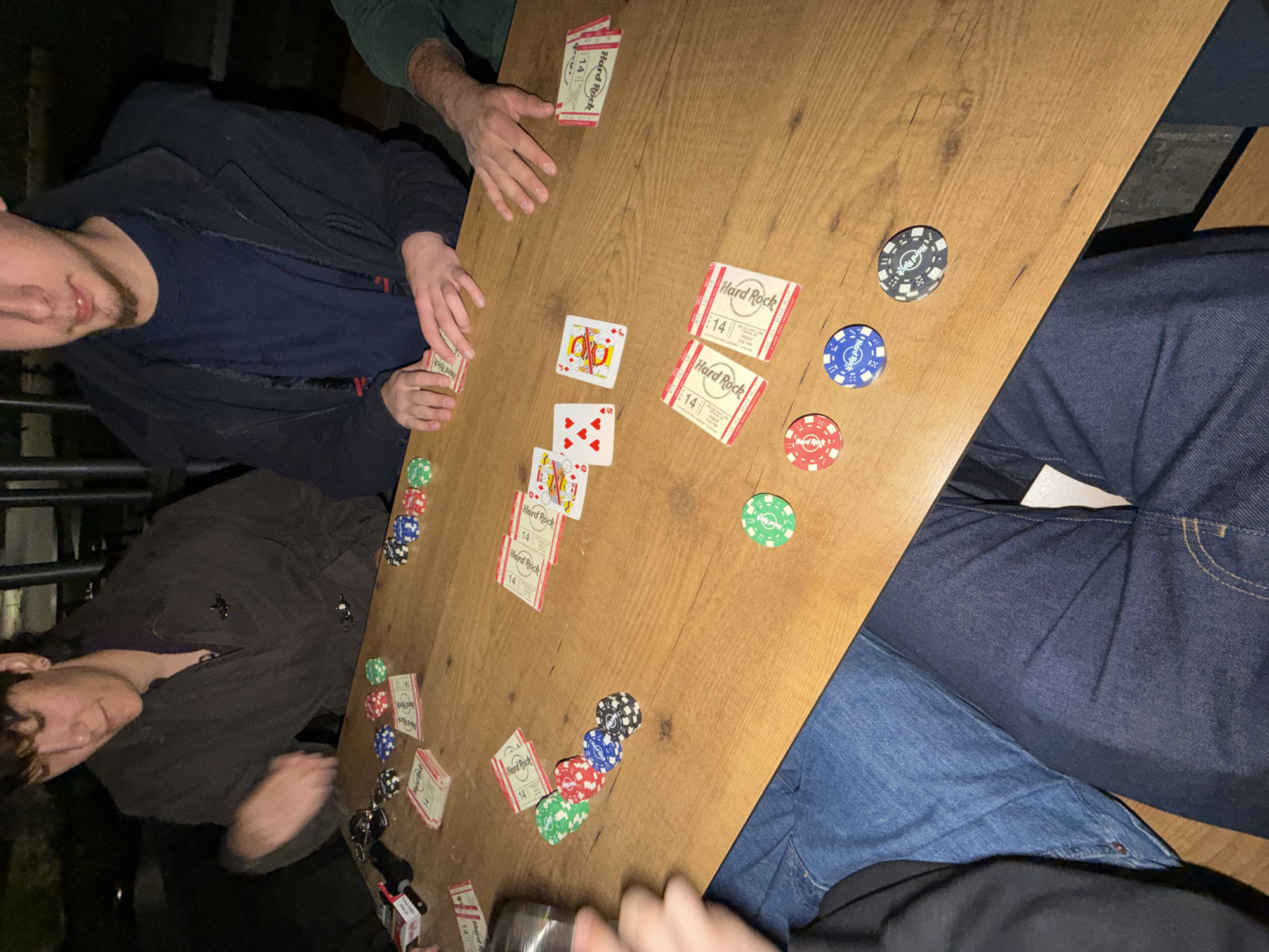 The image depicts a group of people engaged in a card game on a wooden table. Several playing cards and poker chips are scattered on the table. Notably, some of the cards are branded with the "Hard Rock" logo and the number "14." The presence of these Hard Rock branded items suggests a possible connection to the Hard Rock Cafe chain. However, this cafe is not located in Vila Real, Portugal, so this setting is likely a different location. There are multiple individuals partially visible around the table, suggesting a social gathering centered around this card game. The lighting appears dim, indicating that it may be nighttime or an indoor setting with subdued illumination.