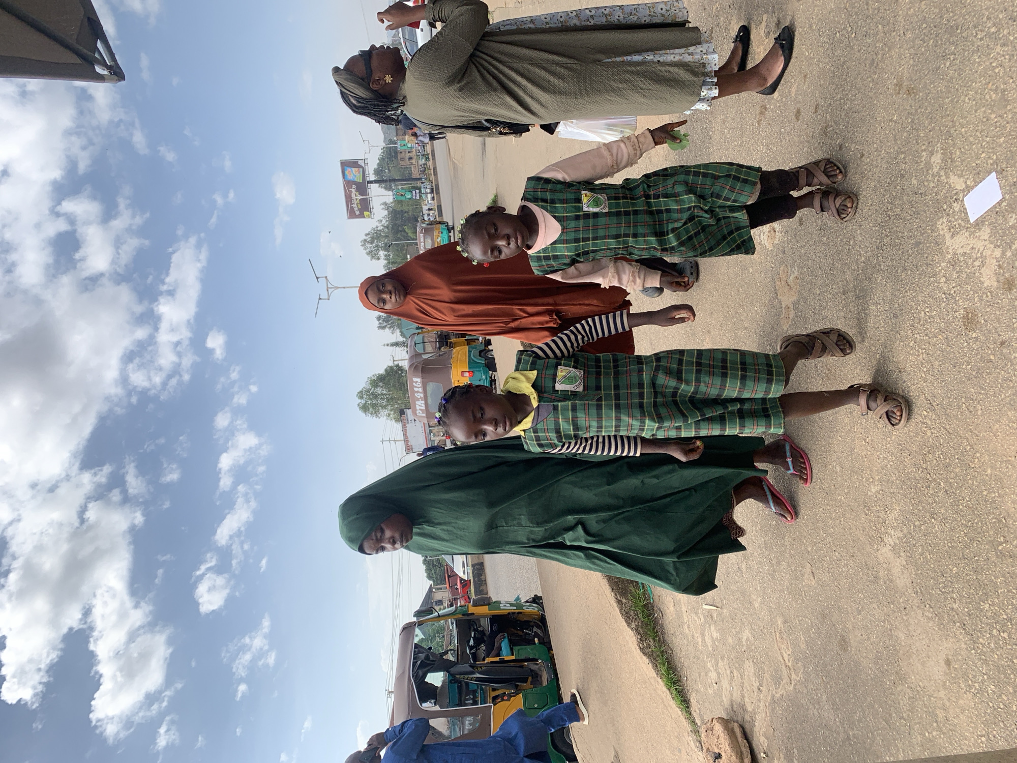 An outdoor, medium-shot photograph captures a group of people walking on a paved street in Jos, Nigeria. The sky is a pale blue with scattered white clouds. In the foreground, two young girls in matching green plaid school uniforms walk side-by-side. The girl on the left wears a green and black striped long-sleeved shirt under her uniform, while the girl on the right wears a light pink long-sleeved shirt. Both have small flowers in their hair.
Behind them, a woman in a long, flowing, dark green hijab walks with her head slightly bowed. To her right, another woman in a rust-colored hijab stands slightly behind the first woman. Further to the right, a woman wearing sunglasses and a green headscarf and dress walks away from the camera, holding her hand up to her face.
In the background, a yellow and green auto-rickshaw is stopped at the side of the road, with a driver in the driver's seat. A man in a blue outfit stands near the rickshaw, talking on a cell phone. Traffic signs, streetlights, and billboards are visible in the distance, indicating an urban environment. The street is lined with trees and some sparse vegetation. The overall scene depicts a common street view in Jos.