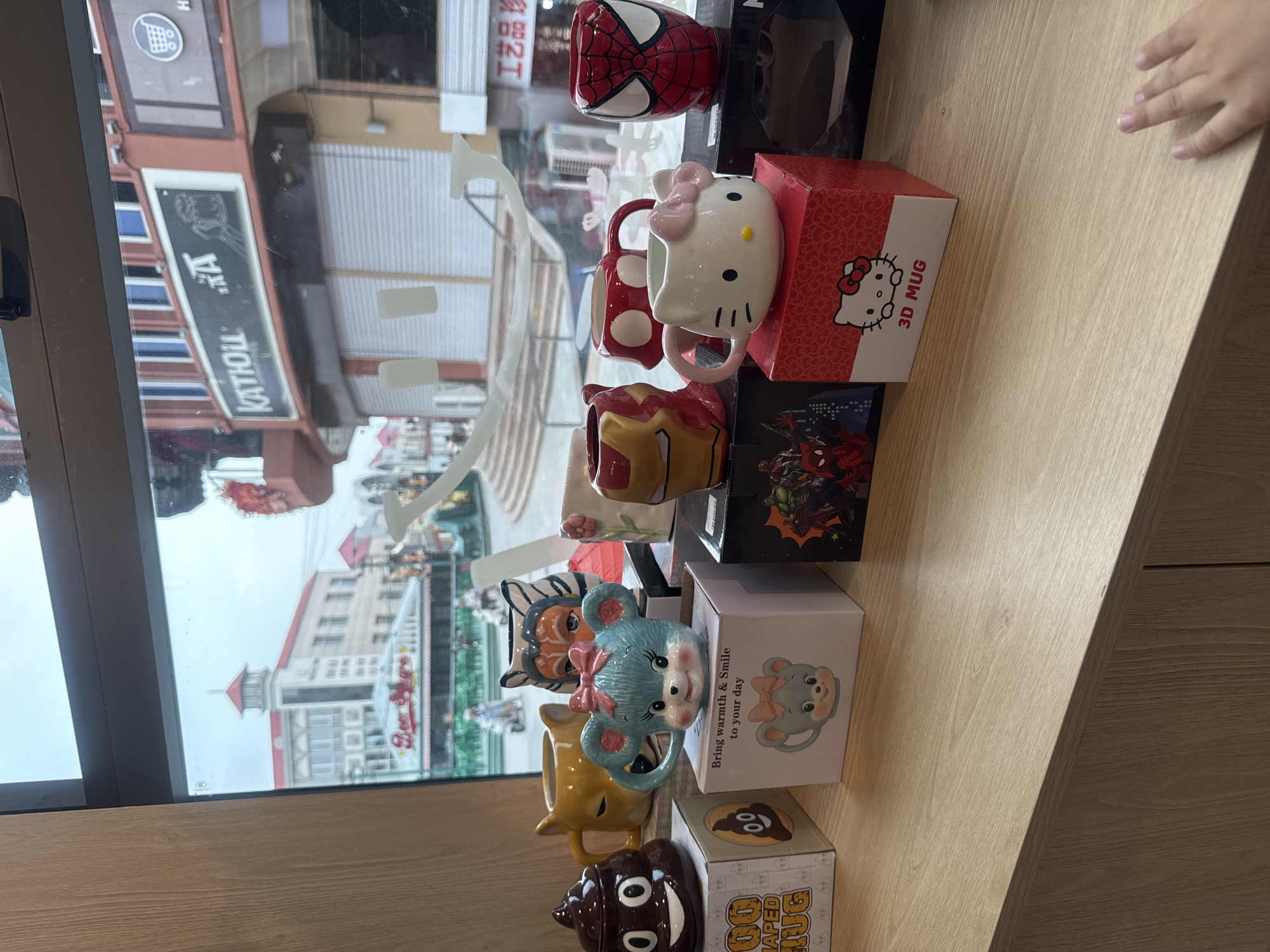 In Qinhuangdao, China, a collection of novelty mugs sits on a wooden counter in front of a window. The mugs, each shaped like a cartoon character or item, are displayed on their respective boxes. Visible mugs include a brown poop-shaped mug, a yellow lion-shaped mug, a blue mouse-shaped mug with a pink bow, an Iron Man helmet mug, a red mug with white polka dots, a Spiderman mug, and a Hello Kitty mug. The window overlooks a street scene with buildings, shops, and people, suggesting a commercial area. The overall impression is of a retail display in a store.