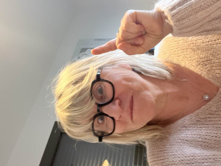 The image displays an indoor scene featuring an adult individual with blonde, shoulder-length hair and bangs, centered in the frame. The individual wears dark, thick-framed glasses, through which their eyes are wide open. Their mouth is closed, with lips slightly pursed, conveying a playful expression. Dimples are visible on the cheeks. They are dressed in a light-colored, possibly beige or light pink, knitted V-neck sweater. A silver-colored chain necklace with a circular, possibly pearl-centered pendant, hangs around their neck. The individual's right hand is raised, with the index finger extended vertically upwards; a ring is visible on the ring finger. The background consists of a plain, light-colored wall. To the far left, a dark, horizontal Venetian blind is partially visible, and a subtle light fixture is implied above it. To the right, a vertical dark line suggests a door frame or edge of a cabinet, with faint horizontal striations on it. The image itself provides no discernible features or contextual clues to confirm the location as Chambœuf, France.