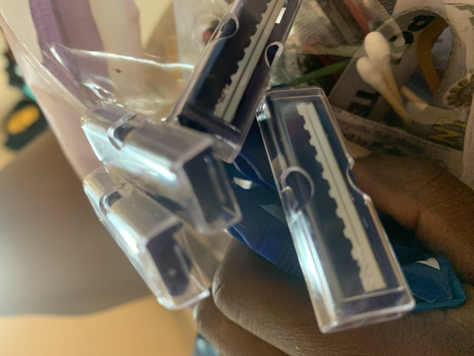 The image shows a close-up of three disposable razors with blue handles, each in a clear plastic protective cover. They are clustered together, with one razor angled slightly upwards. Behind the razors, a blurred background suggests an indoor setting with various items, including what appear to be cotton swabs and fabric. The lighting is bright, casting reflections on the plastic covers of the razors. The image is likely taken by a person, as a hand is visible in the lower left corner, holding some of the objects. The overall impression is one of personal grooming items.