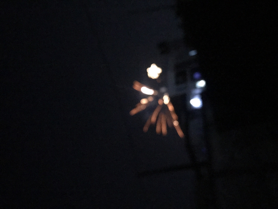 In a dark night scene, a blurry burst of light resembling fireworks appears. The light radiates outwards, featuring a central, star-shaped glow with smaller, linear trails extending downwards and outwards. The image is out of focus, lending to a hazy quality. Adjacent to the burst are additional blurred points of light, possibly streetlights or other distant sources. The surrounding area is enveloped in darkness.
