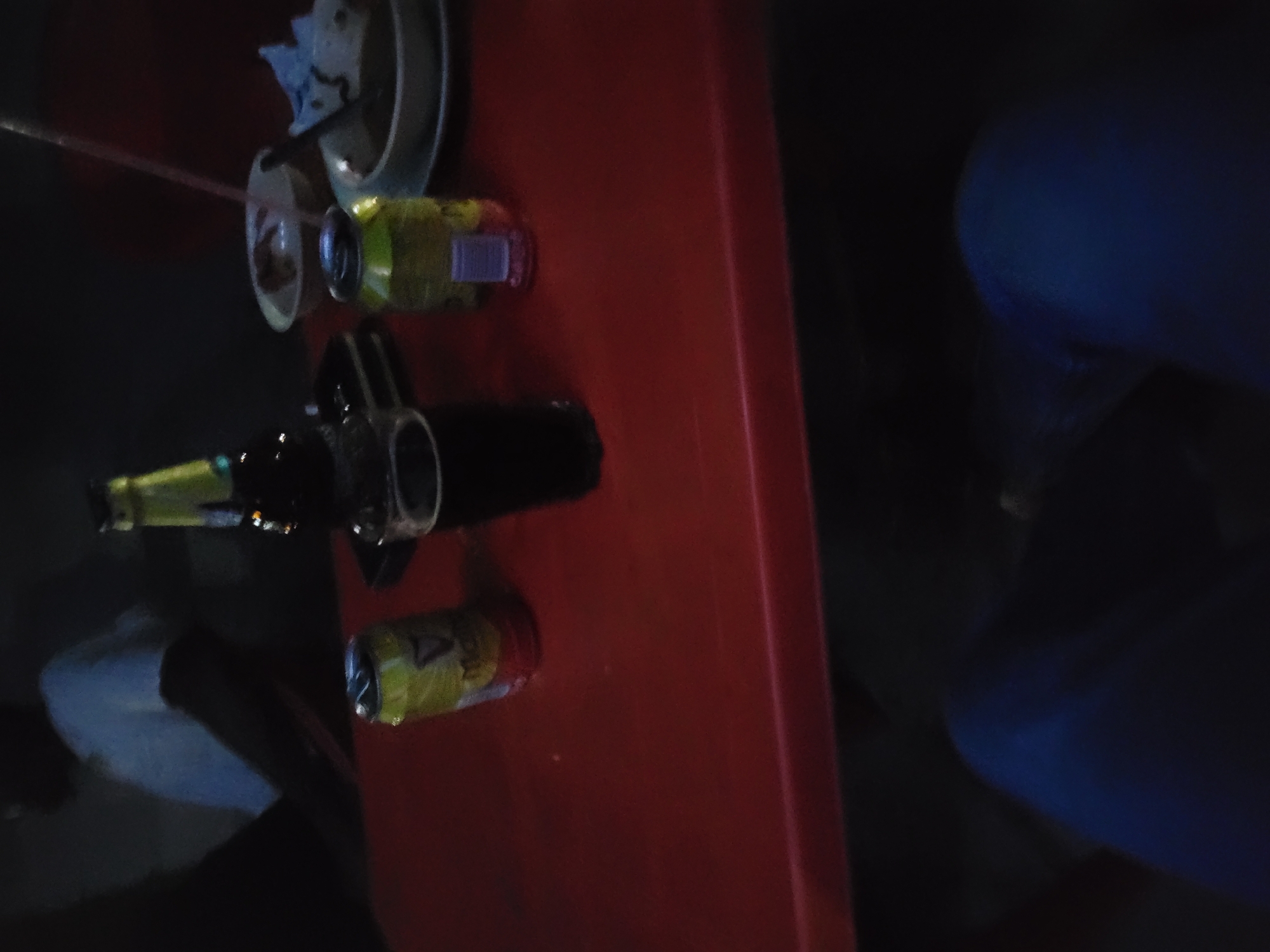 The image depicts a dimly lit scene at a location in Keffi, Nigeria. A red rectangular table occupies the foreground and midground. Underneath the table, a person's legs, clad in blue trousers, are visible. In the background, to the upper left, a person is partially visible from the chest upwards, wearing a light-colored top, with their face obscured by darkness.
On the red table, multiple items are arranged:
- To the left, an open yellow aluminum can, bearing a partial "GUINNESS" logo, is present.
- Adjacent to it, a clear glass filled with a dark liquid (consistent with stout or dark beer) is visible.
- Behind the glass and can, a brown glass bottle with a yellow and green label and cap is positioned.
- Further to the right, a second yellow aluminum can, similar to the first, is also open.
- Between these two cans, a small, blurry, purple object, possibly a miniature bottle or container, rests on the table.
- On the right side of the table, two white bowls are present. The rear bowl contains a light-colored substance, and a pink drinking straw is inserted into it or nearby. The front bowl contains a darker, viscous substance, possibly food, with a crumpled light-colored napkin partially submerged or resting on its edge.
The arrangement of beverages and food suggests a casual social gathering or meal in a low-light environment, likely an evening or nighttime setting.