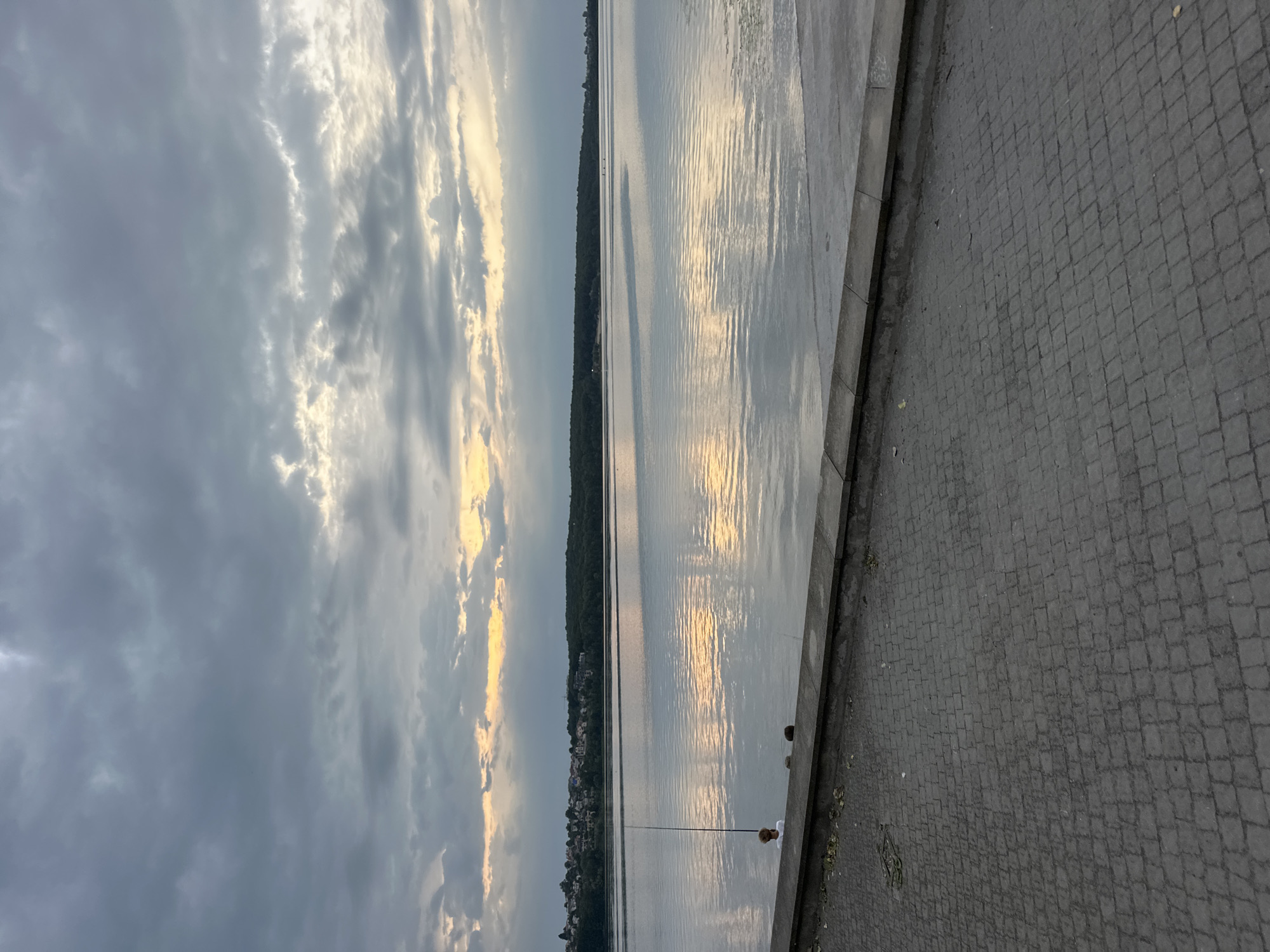 The image depicts a wide, calm body of water, likely Ternopil Lake, under a dramatic, cloudy sky. The water reflects the muted colors of the sunset and the overcast sky, creating a serene and somewhat somber atmosphere. A paved walkway with cobblestone-like patterns lines the foreground, leading to a concrete embankment. On the embankment, a person is visible, appearing to be fishing with a long rod. Another person is seated further down the embankment. The opposite shore is lined with a dense forest and a cluster of buildings, suggesting a town or city bordering the lake. The overall scene captures a quiet moment by the water, possibly during an evening or early morning, as indicated by the lighting and cloud cover.
