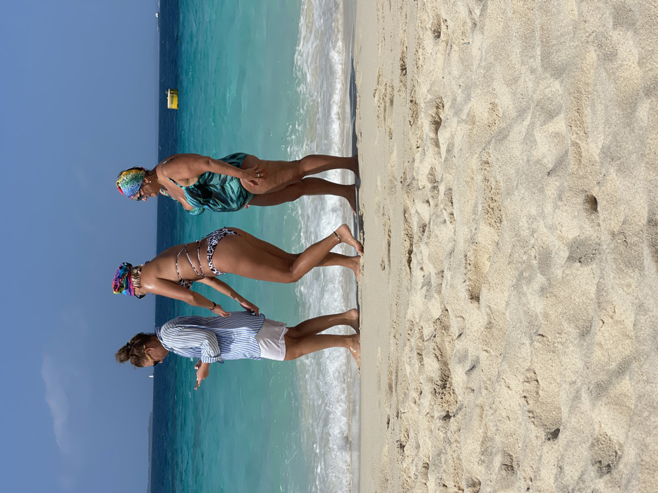 The image displays three adult females walking barefoot along the waterline of a sandy beach in Zanzibar, Tanzania, under a clear blue sky. The foreground consists of light-colored sand marked with minor depressions and undulations.
From left to right:
1. A woman with dark hair pulled into a high bun wears sunglasses, a light blue and white striped long-sleeved button-up shirt, and white shorts. She is looking down, holding a smartphone in both hands. Her bare feet are partially submerged in the shallow water.
2. A second woman, with blonde hair covered by a multi-colored patterned headscarf, wears a leopard-print two-piece string bikini featuring multiple criss-cross straps on her torso. She has a dark anklet on her right ankle. Her right hand is extended backward, lightly grasping the lower back of the first woman's shirt. Her bare feet are also in the shallow water.
3. A third woman, appearing older, wears sunglasses and a multi-colored patterned headscarf similar to the second woman's. She is dressed in a teal one-piece swimsuit with a dark green leaf-like pattern. Her bare feet are submerged in the water.
All three individuals are positioned facing generally towards the right side of the frame, walking parallel to the shoreline. The ocean water is clear and turquoise blue, with small white waves gently breaking at their feet. In the background, the vast blue ocean extends to a distant horizon. A faint landmass or island is visible on the far left horizon, with a small white object, possibly a sailboat or marker, positioned near it. A yellow rectangular buoy floats on the water further out towards the right side of the frame. The sky above is clear and bright blue, indicative of a sunny day.