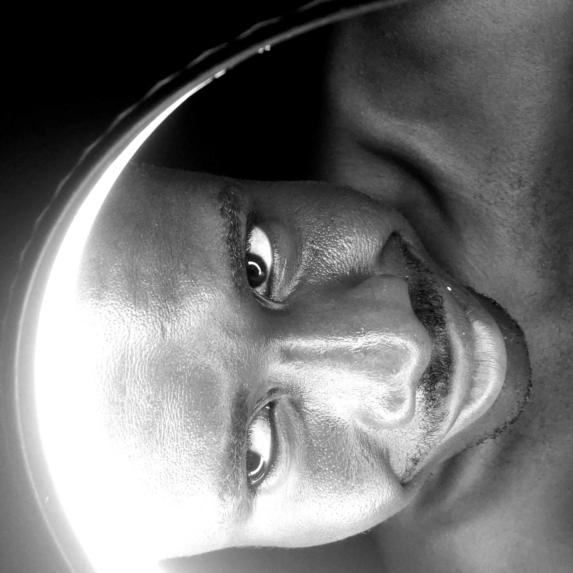 A striking black and white close-up portrait captures a dark-skinned person, likely male, looking directly into the camera. The image is taken in Narayi, Nigeria. The subject's face is partially illuminated by a prominent, circular ring light, which creates a bright, reflective highlight down the left side of the face (when viewed upright). The rest of the scene, including the background and part of the subject's body, is enveloped in deep shadow, emphasizing the dramatic lighting and the subject's features.
The subject's eyes are wide open, intently fixed on the viewer, with distinct crescent-shaped reflections of the ring light visible in both pupils. Visible details include stubble or short facial hair on the upper lip and chin, as well as distinct skin texture on the forehead and cheeks, accentuated by the harsh light. The angle of the shot appears to be from below, with the subject's head tilted. Only the head, neck, and a portion of the upper shoulder/chest, which appears bare, are visible. There is no visible text in the image. The overall impression is one of direct engagement and intensity due to the strong gaze and high-contrast, artistic lighting.