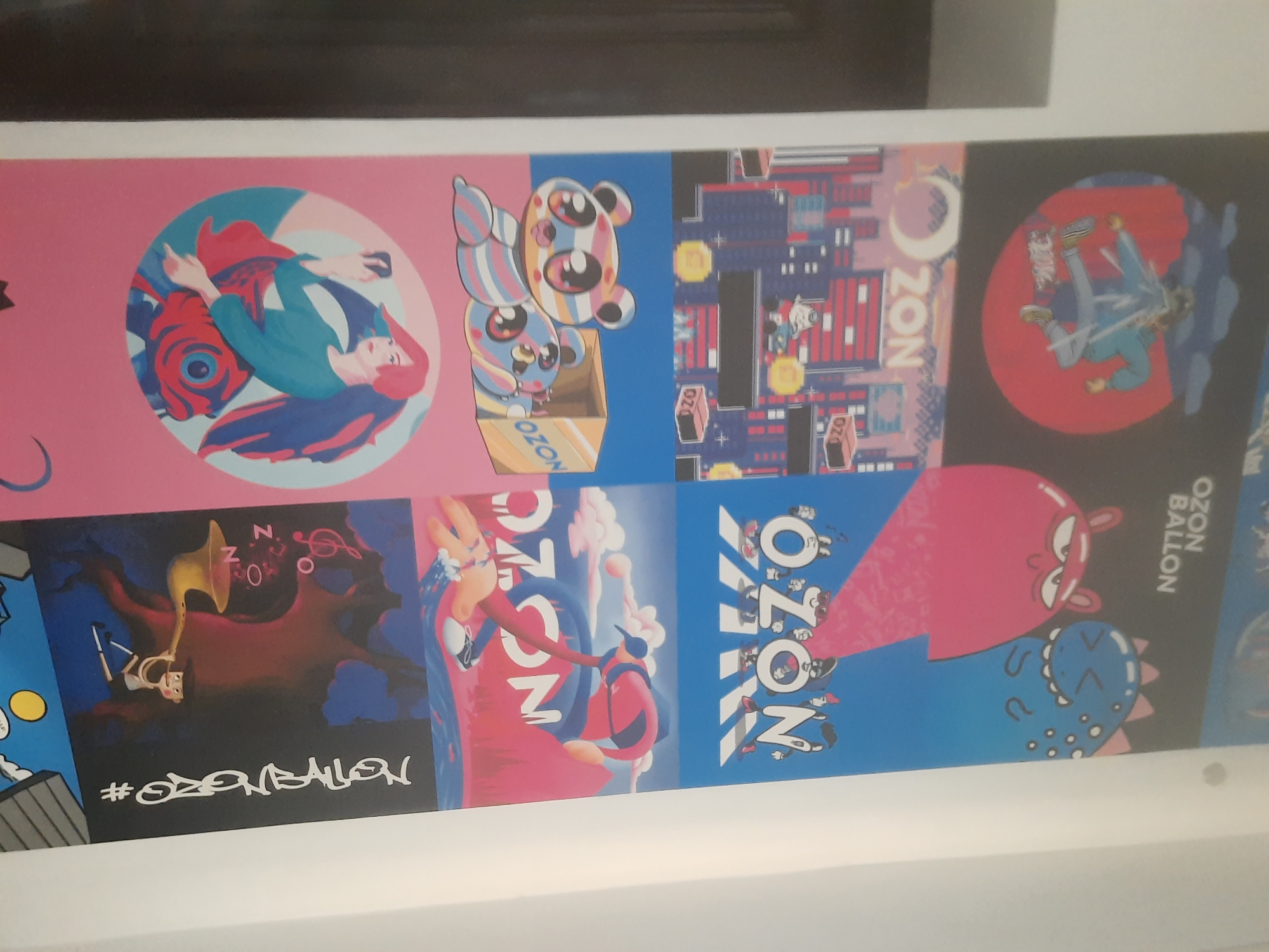 The image shows a vertical poster with multiple illustrated panels, displayed on a white wall in what appears to be an indoor setting, possibly a store or gallery. The poster's content is a series of colorful, cartoonish graphics with a consistent theme, likely promoting a brand or event.
The top panel depicts a person in a blue hoodie and pants, mid-air, with a red circle as a background. The text "OZON BALLON" is visible in the upper right section of this panel.
Below this, two cartoon monsters are featured against a blue background. The left monster is pink with large eyes, and the right is blue with teeth and polka dots.
The next panel shows a cityscape with pink and blue buildings and a yellow moon. The word "OZON" is prominently displayed in large white letters, with small figures in a video game-like setting below.
Following this, two striped panda-like characters are illustrated on a pink background. One is sitting, and the other is peeking out of a cardboard box labeled "OZON."
The fifth panel features a stylized depiction of a person with red hair and a blue shirt, holding a phone and appearing to take a selfie, with a large red and blue fish motif.
The bottom panel shows a dark, silhouetted forest scene with musical notes and the word "OZON" repeated. A small figure is playing a golden horn. To the right of this panel, the hashtag "#OGANANGRO" is written vertically in white graffiti-style lettering.
There is no indication of the location being in Tashkent, Uzbekistan. The image is a close-up of a poster, and the surrounding environment is minimalist, focusing on the white wall.