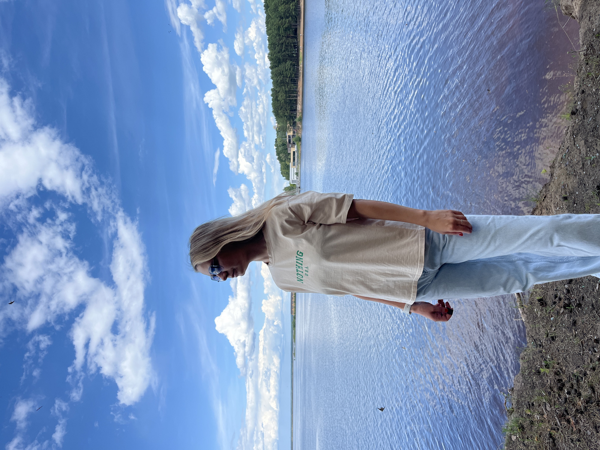 A young woman with blonde hair and sunglasses stands on the shore of a body of water. She is wearing a light-colored t-shirt with green text that reads "NOTHING U.S.A." and light blue jeans. The sky is a bright blue with fluffy white clouds. A line of trees is visible across the water, and there are some buildings nestled among the trees. The water is calm, with ripples indicating a gentle breeze. The ground on the right side of the image is covered in dry dirt and sparse vegetation. The image appears to be taken during the daytime, with bright sunlight casting shadows. The woman is looking down, and her posture suggests a relaxed or contemplative mood. The overall scene evokes a sense of tranquility and natural beauty.