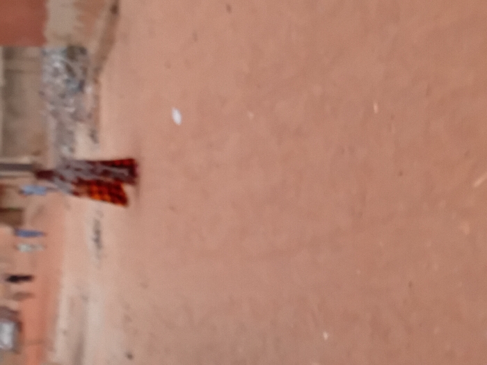 The scene depicts an unpaved, reddish-brown earthen road in Minna, Nigeria, under daylight conditions. In the mid-ground, a single individual is walking away from the camera, centered in the frame. This person wears a long, patterned garment exhibiting colors such as red, orange, and dark hues. Further down the road, in the background, several additional blurry human figures are visible, some appearing to be walking. To the far left in the background, a highly indistinct vehicle is present. On the right side of the road in the mid-background, a pile of assorted debris is observed adjacent to a light-colored wall or building. The foreground and mid-ground of the road surface show small scattered debris, including a distinct white object on the right side. The overall environment suggests a residential or semi-urban area with structures constructed from reddish-brown materials.