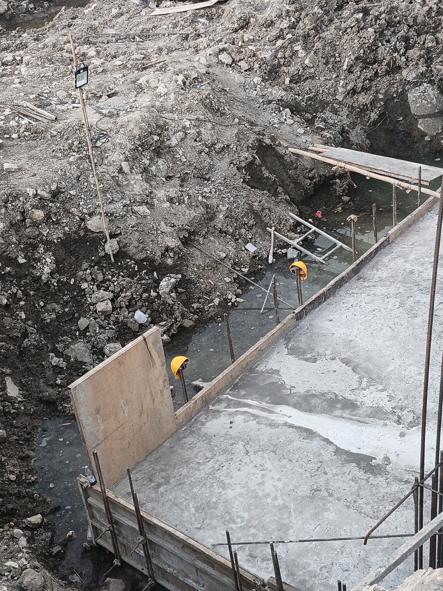 An aerial view of a construction site in Planggu, Indonesia, shows a partially poured concrete foundation surrounded by excavated earth and debris. A wooden formwork defines the perimeter of the concrete slab, with rebar visible protruding from it. Two orange safety helmets rest on vertical poles within the excavated area, one of which is partially submerged in muddy water. A tall wooden pole with a light fixture is planted in the surrounding dirt pile. The ground is a mix of mud, rocks, and some wooden planks. The scene suggests ongoing construction work, possibly for a building or infrastructure project.