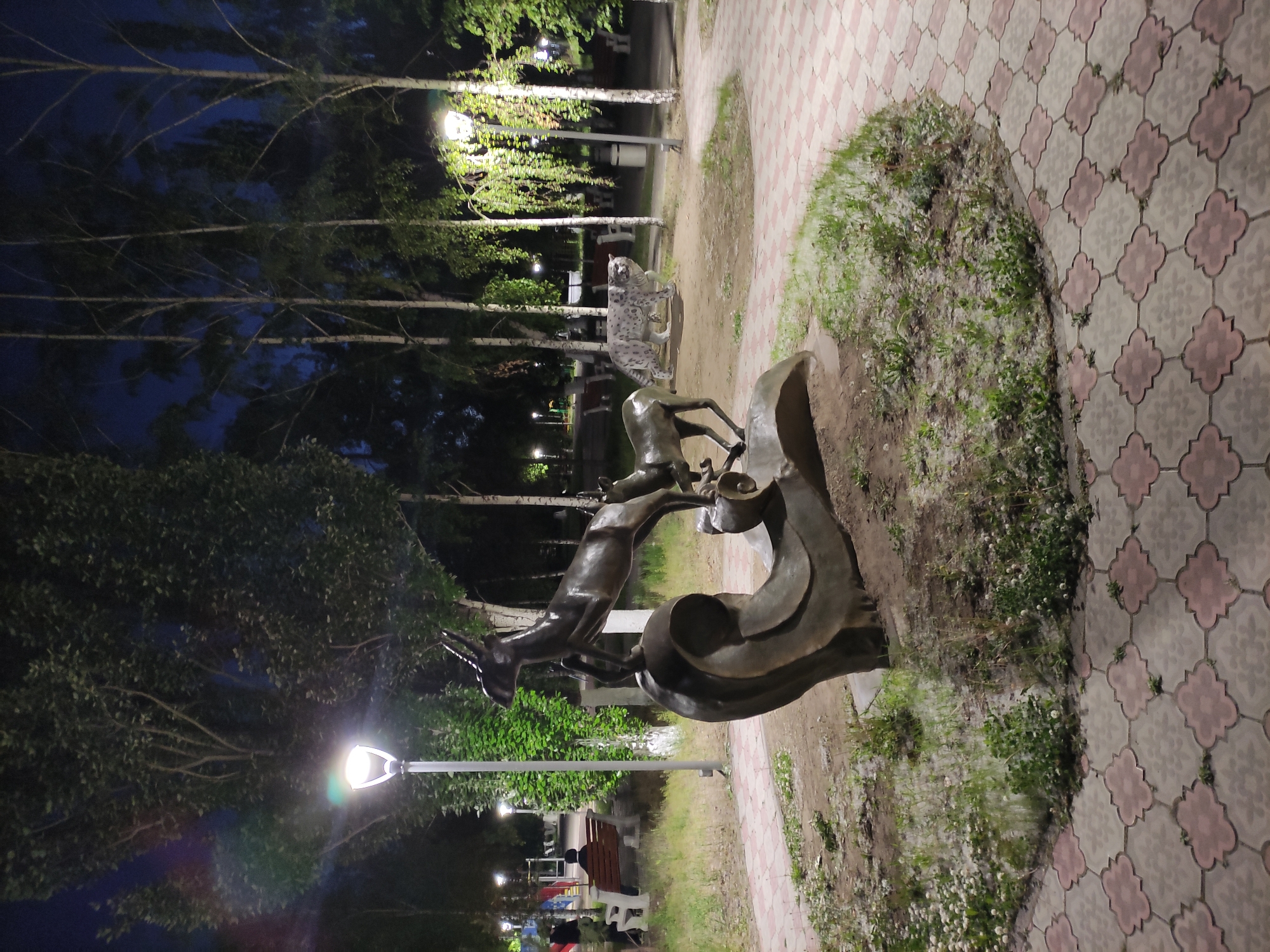 The image shows a bronze statue of two gazelles leaping, located in a park in Pavlodar, Kazakhstan. Behind the gazelle statue is a concrete sculpture of a snow leopard, situated among birch trees under streetlights. A tiled walkway borders the statue. In the background, park benches and recreational equipment are visible, indicating a public park setting. The photograph was taken at night.