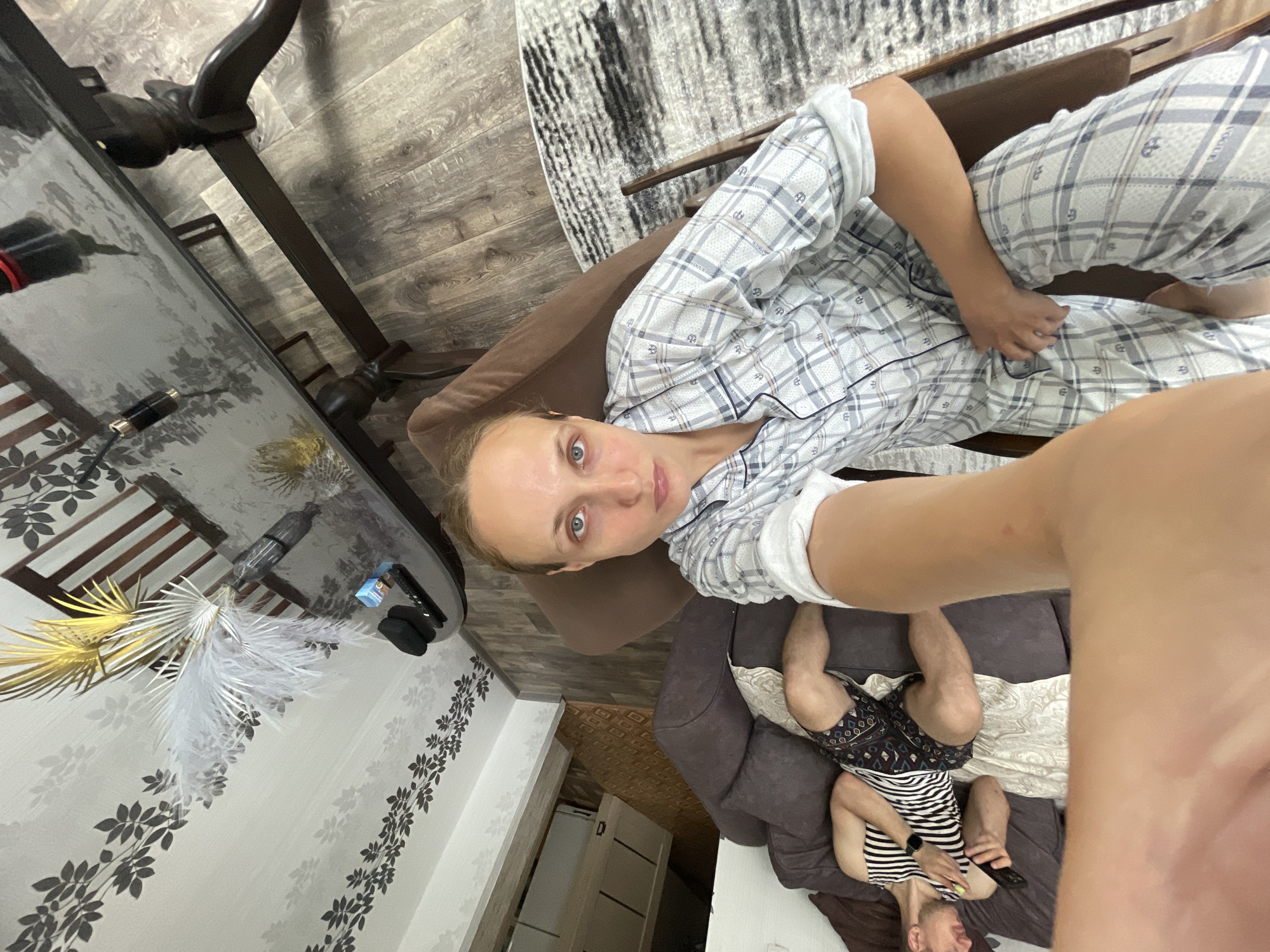 A woman is taking a selfie in a living room. She is sitting in a brown chair and wearing pajamas. A man is lying on a sofa next to her, also wearing pajamas. The room has wooden floors and a glass-top table. There is also a rug and a vase with decorative feathers on the table. The image does not contain any information indicating the location is in Sharkan, Russia.