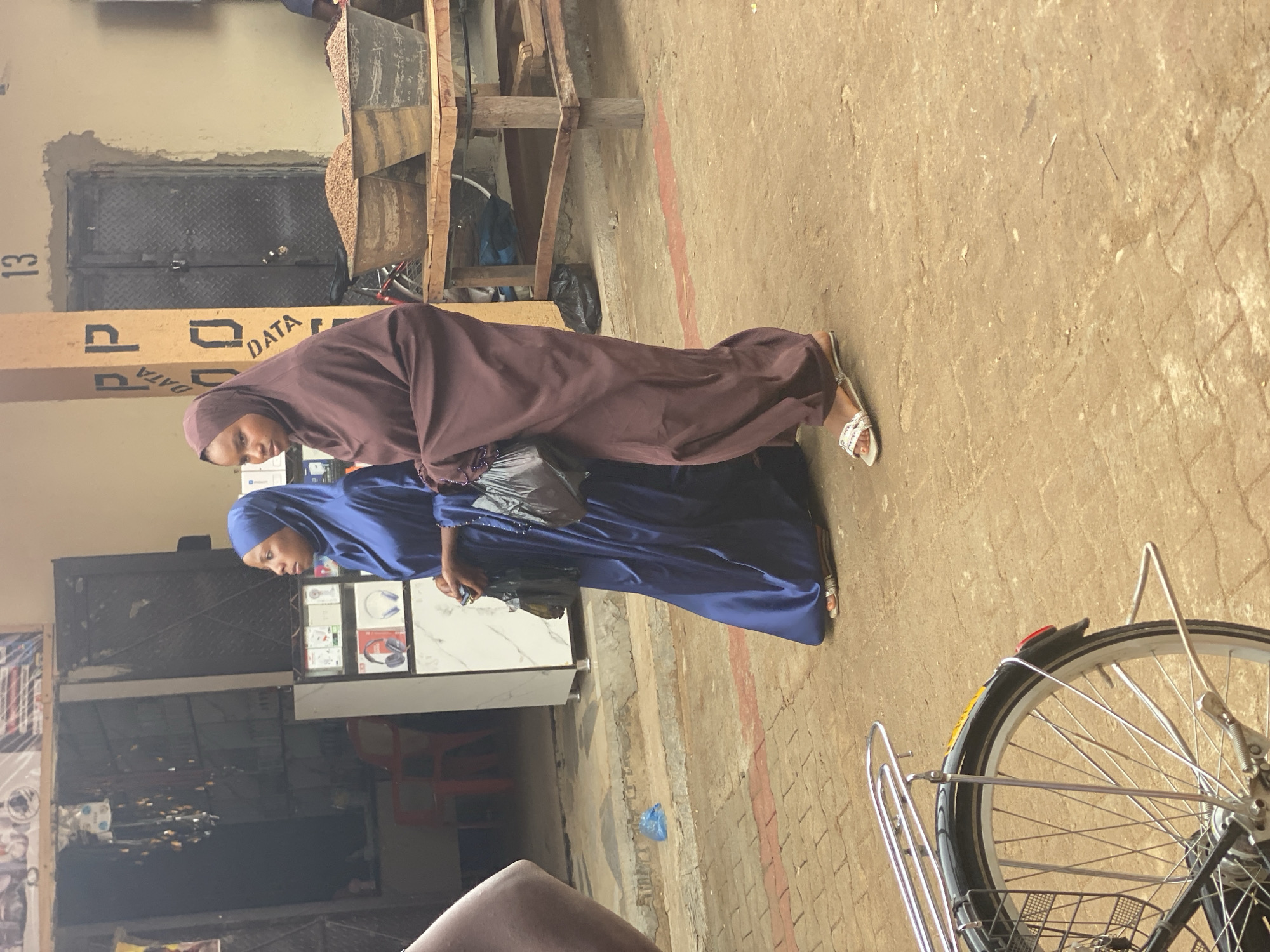 Two women wearing hijabs are standing next to each other on a paved area near what appears to be a storefront. The woman in the front is wearing a blue hijab and long dress, while the woman behind her is wearing a brown hijab and dress. They are standing close to a bicycle, with the back wheel visible in the foreground. Behind them are buildings and a wooden structure holding what looks like a pile of nuts. There is a sign with the word "DATA" visible. The scene is outdoors and appears to be a market area in Kafanchan B, Nigeria.