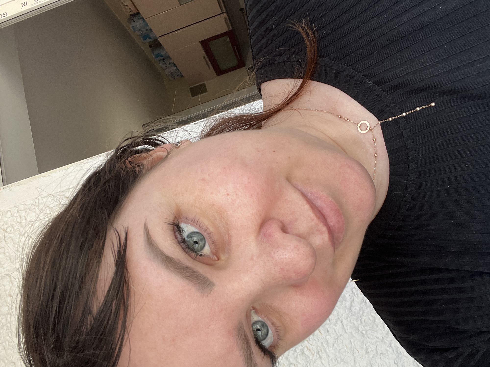 A close-up, angled selfie of a woman with gray-blue eyes, light skin, and brown hair. She is wearing a black ribbed top and a delicate gold necklace with a circular pendant. The background is blurry but appears to include a white textured surface and a partial view of cabinets or storage units. The location in the city of Noviy, Russia, cannot be determined from the visual data provided.