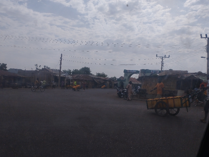 The image depicts a street in Kumo, Nigeria (10.049, 11.2), with several people and vehicles, including a man pushing a handcart with a yellow container in the foreground. The road is paved and has a few small buildings on either side, some with thatched roofs. There are overhead power lines and numerous trees in the background. The sky is cloudy with a few patches of blue visible.  The scene appears to be a commercial area with numerous people engaged in various activities.  A group of men are walking towards the left side of the frame, while a man is sitting on a motorcycle on the right side of the frame. A few other people are walking in the distance. The scene is located in the Kumo area of Nigeria.