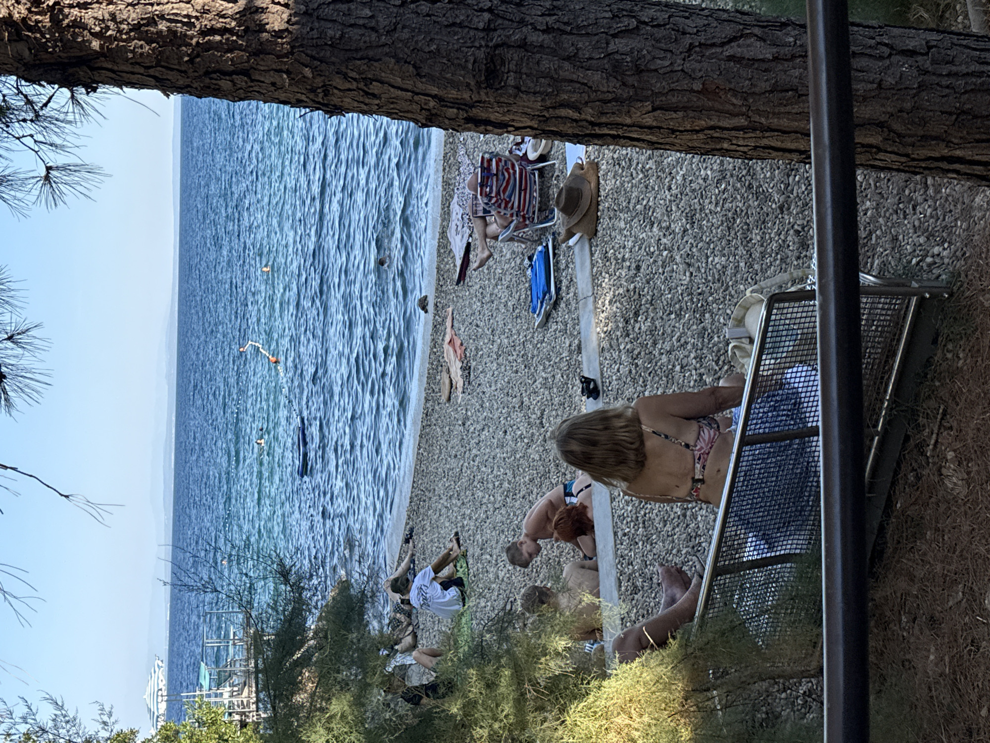 The image depicts a sunny day at a pebble beach in Opatija, Croatia. The tranquil blue sea stretches to the horizon, dotted with a few swimmers and a blue inflatable mat. The beach is populated by several individuals relaxing on towels and chairs. In the foreground, a woman in a bikini sits on a bench facing away from the camera, with a man in swimwear to her left. Behind them, a boy in swim trunks is interacting with another person. Further up the beach, various individuals are sunbathing, some lying on towels, others seated in beach chairs. A tree trunk on the right side of the frame partially obscures the view of the beach. The presence of beach umbrellas and a distant boat suggests a typical summer day at a coastal resort.