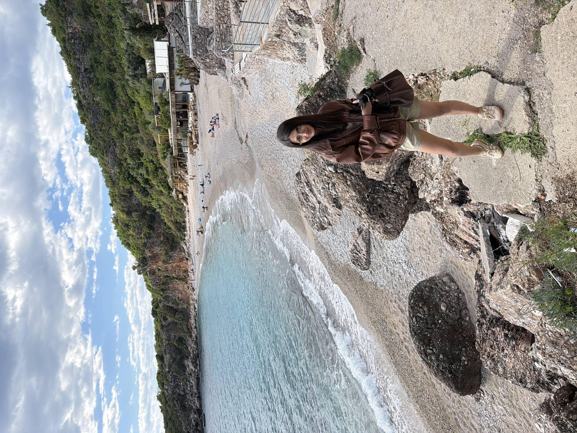 A young woman stands on a rocky outcrop, overlooking a beach near Drobnići, Montenegro. The scene includes a beach with clear, turquoise water, a rocky cliff covered in green vegetation, and a partly cloudy sky. The woman is wearing a brown leather jacket, light-colored shorts, and sneakers. She is holding a bag. The beach is sparsely populated with a few people visible in the distance. A building or structure is visible on the beach near the cliff. The image is taken from a high vantage point.
