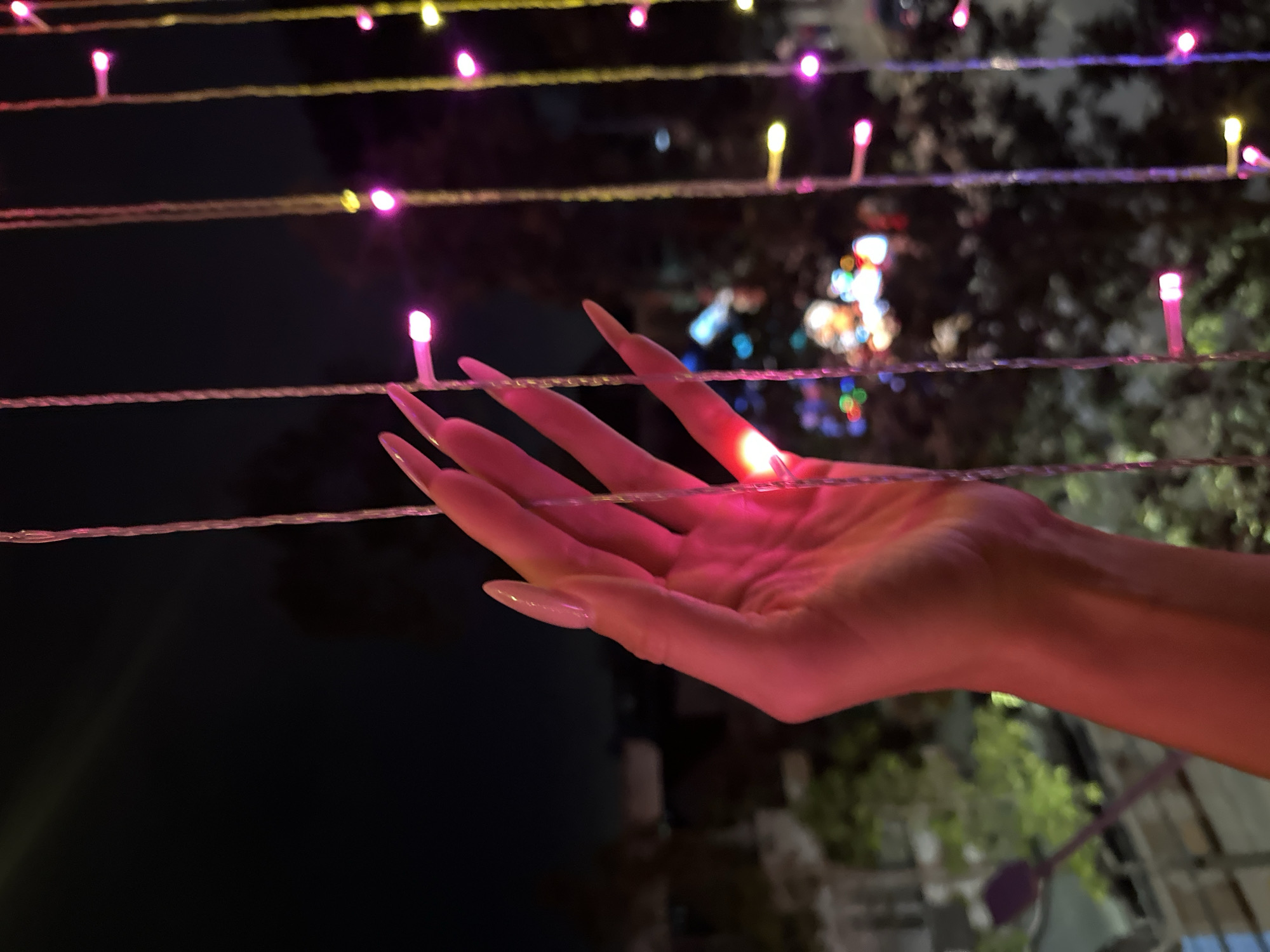 The image shows a hand with long nails, bathed in pink light, positioned between multiple strands of string lights. The lights are of varying colors, including pink and yellow. In the background, out-of-focus lights suggest a city environment, and trees are visible, indicating an outdoor setting. Based on the prompt, this scene is likely in Đồng Nai, Vietnam, at night.