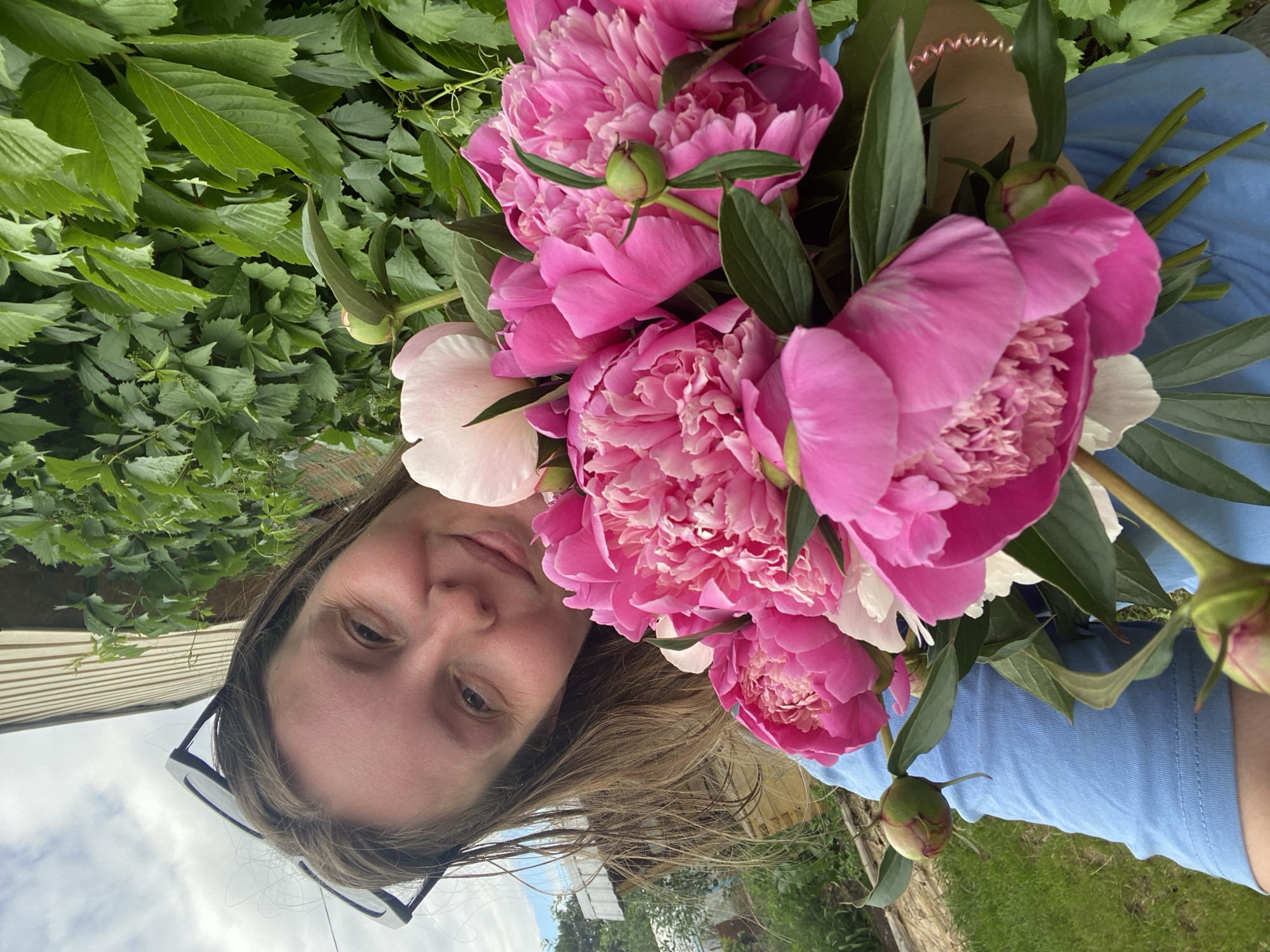 The image shows a young woman holding a bouquet of pink and white peonies, smiling at the camera. She is wearing a blue t-shirt and sunglasses are pushed up on her head. The background features lush green foliage and a building with a light-colored exterior. The scene appears to be outdoors, possibly in a garden or yard, given the presence of grass and the natural setting. It is likely a casual, personal photograph. The context of Chaikovsky, Russia, is not directly evident from visual cues in the image itself.