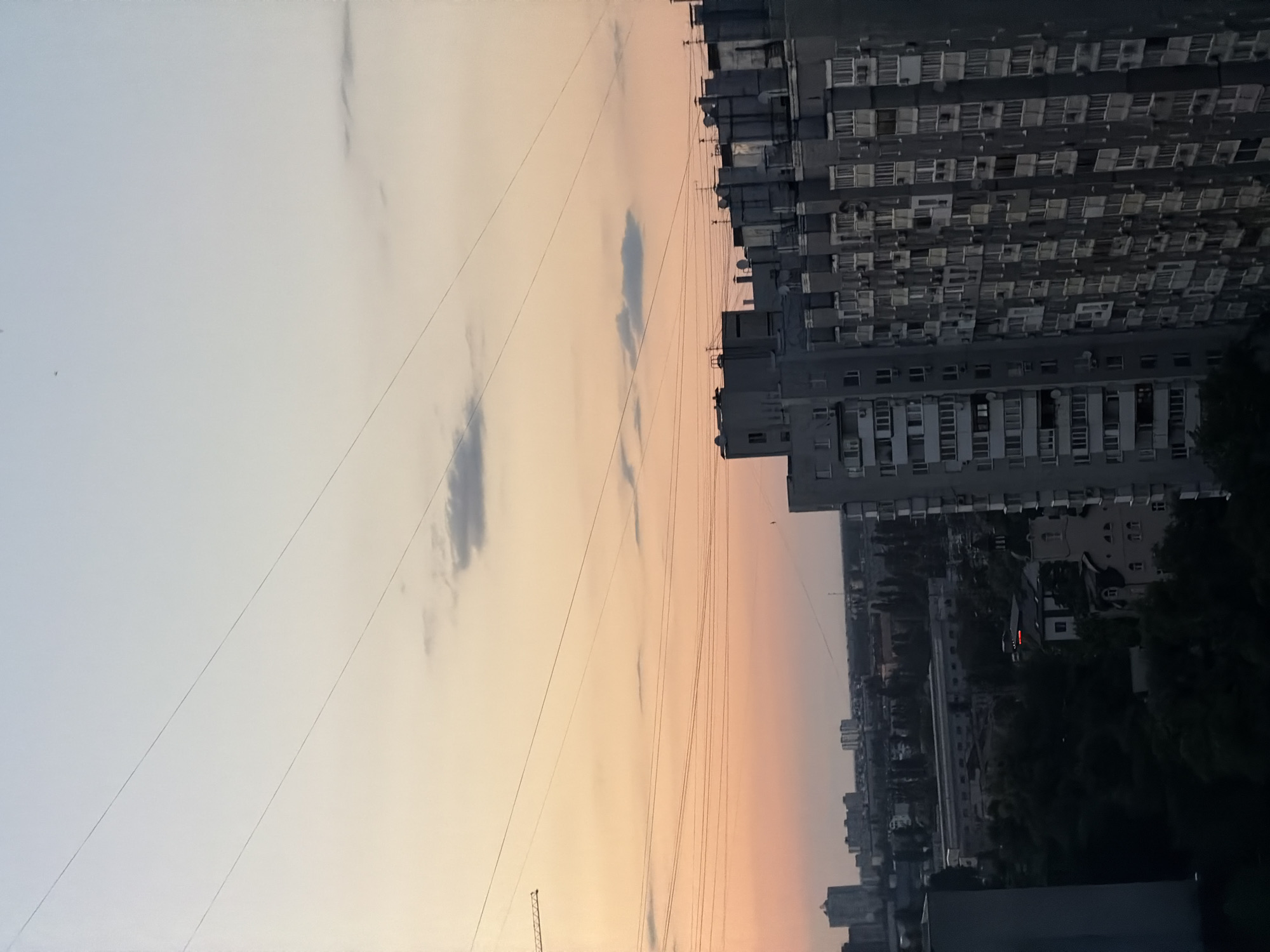The image depicts a city skyline at dusk, likely in Kyiv, Ukraine, characterized by tall residential buildings and a pale, gradient sky transitioning from light blue to orange. Multiple power lines crisscross the frame, some taut and others appearing slack. In the foreground, a large, grey apartment building with numerous balconies dominates the right side. Beyond it, a cluster of similar buildings stretches across the mid-ground, some featuring ventilation systems or air conditioning units on their facades. Further into the background, a denser urban landscape with taller structures is visible, interspersed with trees. A construction crane is partially visible on the far left. The overall impression is one of a densely populated urban environment under a soft, evening sky.
