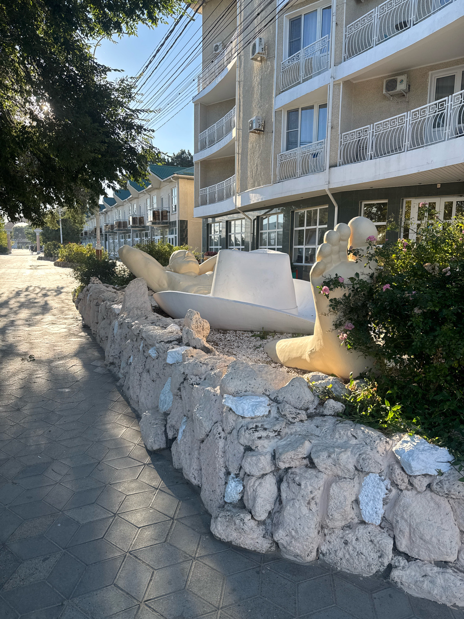 In Anapa, Russia, along a paved walkway lined with a low stone wall, a large, surreal sculpture is displayed. The sculpture depicts a giant, pale yellow foot, resting near what appears to be a large, white hat or bowl. Behind the sculpture, a modern, multi-story building with balconies and air conditioning units is visible, along with a line of similar buildings further down the street. Lush green bushes with small pink flowers partially obscure the sculpture on the right. The walkway is illuminated by sunlight, casting dappled shadows from trees on the left side of the frame. Two people are walking in the distance on the walkway.