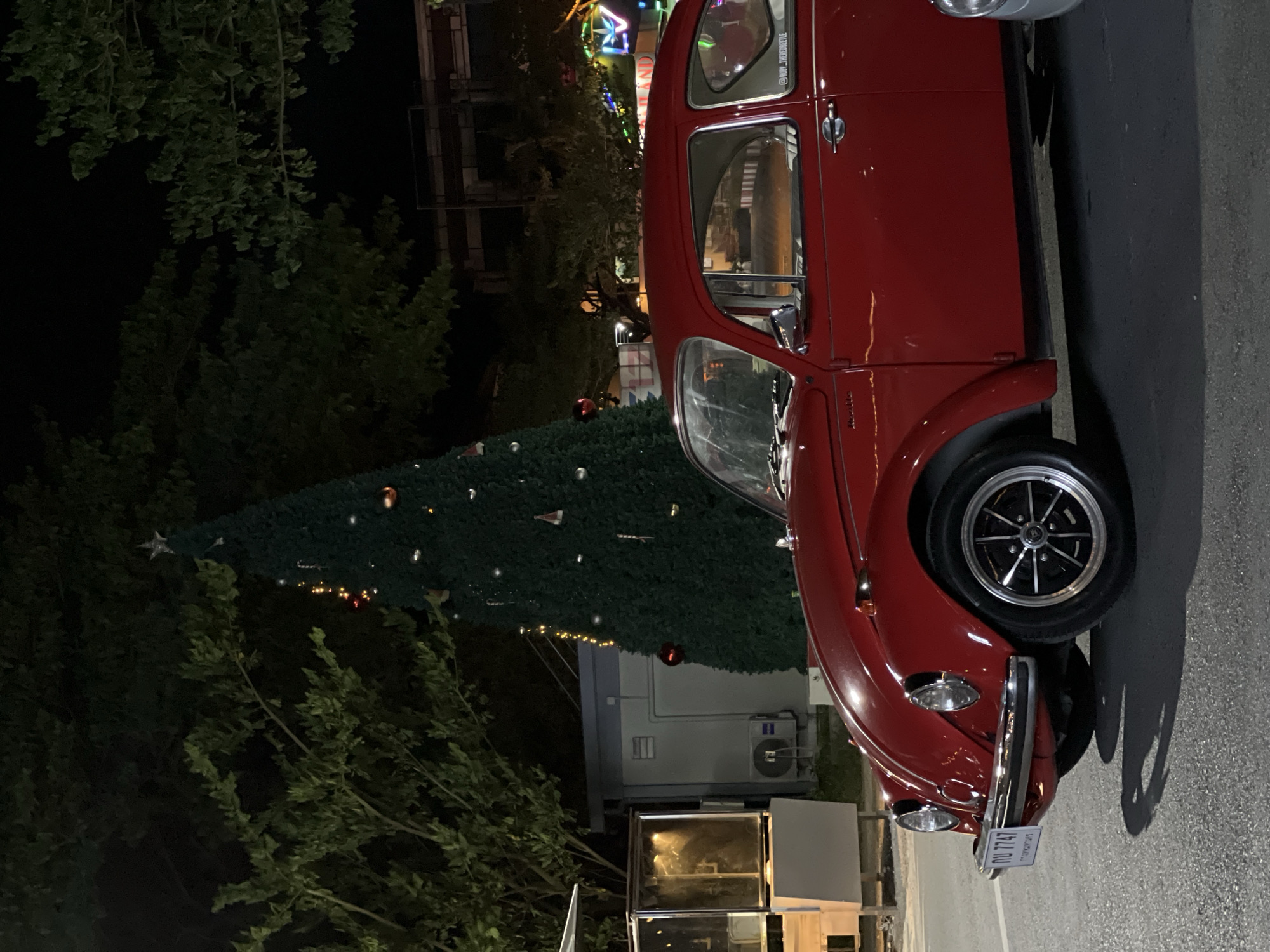 The image captures a vintage red Volkswagen Beetle parked on a paved road at night in Choeng Thale, Thailand. A large, decorated Christmas tree stands to the left of the car, illuminated by small lights and adorned with ornaments. Behind the tree, a grey, modular building with an air conditioning unit is visible. To the far left, a display case with a glass front and lighting is present. In the background, trees with dark foliage are silhouetted against the night sky, and beyond them, streetlights and buildings suggest an urban or commercial setting. The car's license plate, bearing Thai characters and numbers "กบ 7747", is partially visible. The overall scene suggests a festive atmosphere, possibly during the holiday season.