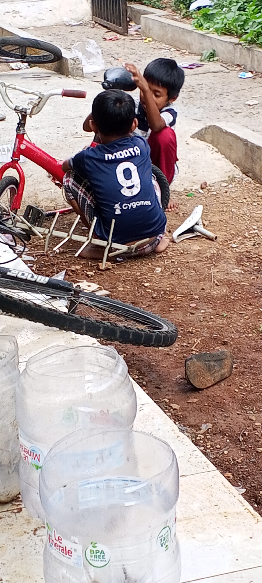 In an outdoor setting in Depok, Indonesia, two young boys are interacting with a dismantled red bicycle. One boy, wearing a blue jersey with the number "9" and "Cygames" printed on it, sits on the ground facing away from the camera, with his back to another boy. His legs are visible beneath a checkered skirt. The second boy, facing the first, kneels behind him, with his hands raised. A pigeon is perched near the first boy's head, and it appears the second boy might be attempting to touch it. The bicycle frame is red, and its components, including wheels and a seat, are scattered around. The ground is covered in dirt and pebbles, with some debris visible. In the foreground, two large plastic bottles, one partially filled with liquid, are placed on a concrete ledge. The background shows more dirt, some greenery, and a wooden fence.