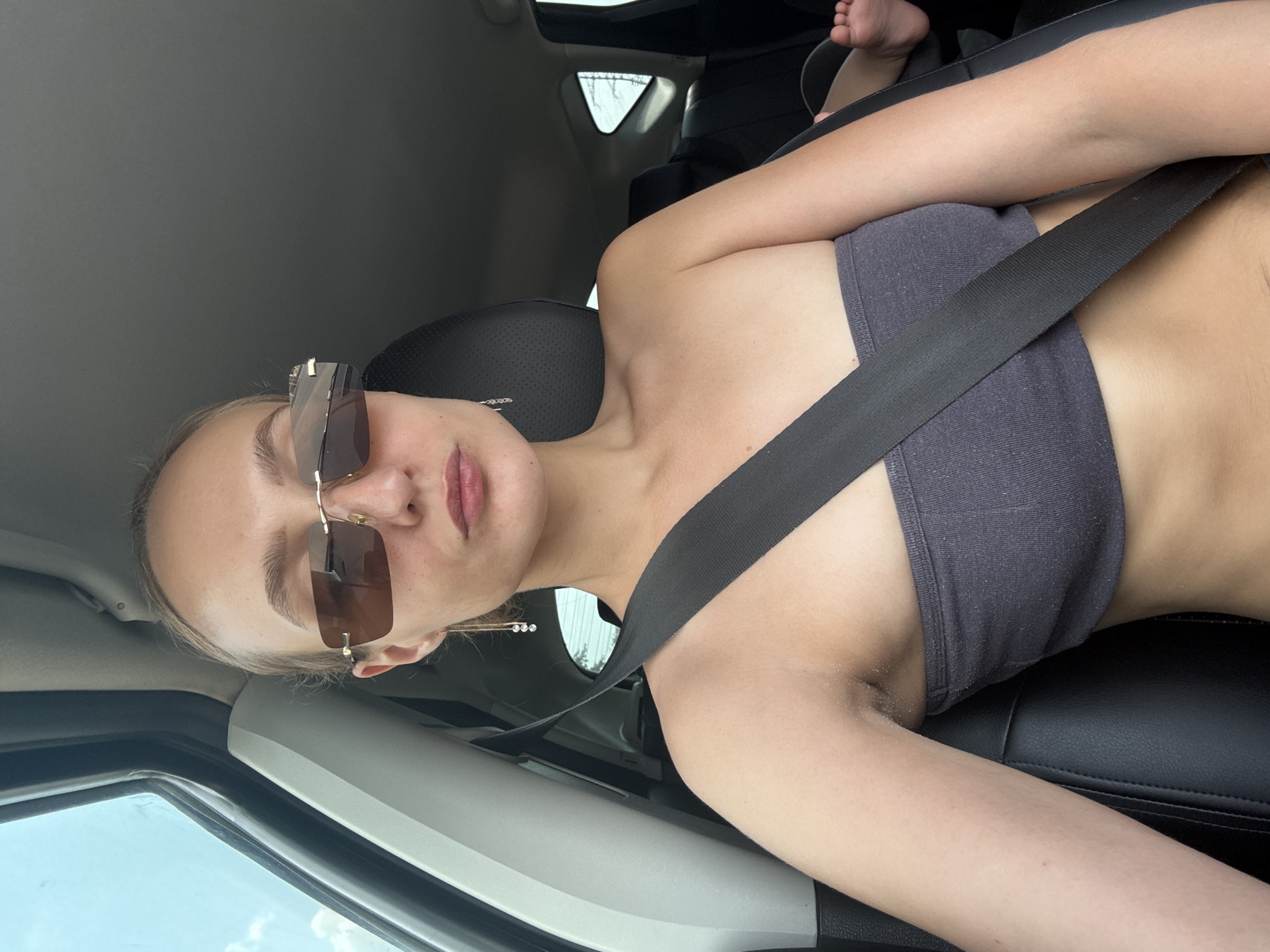 A young woman with blonde hair pulled back is sitting in the passenger seat of a car, wearing a grey strapless top and sunglasses. A black seatbelt is fastened across her body. Her left arm is resting on her lap, and her right arm is bent at the elbow. The interior of the car is visible, with black leather seats and a window on the left showing a cloudy sky. In the background, a small child's foot is visible, suggesting the child is in the back seat. The image was taken in Izhevsk, Russia.