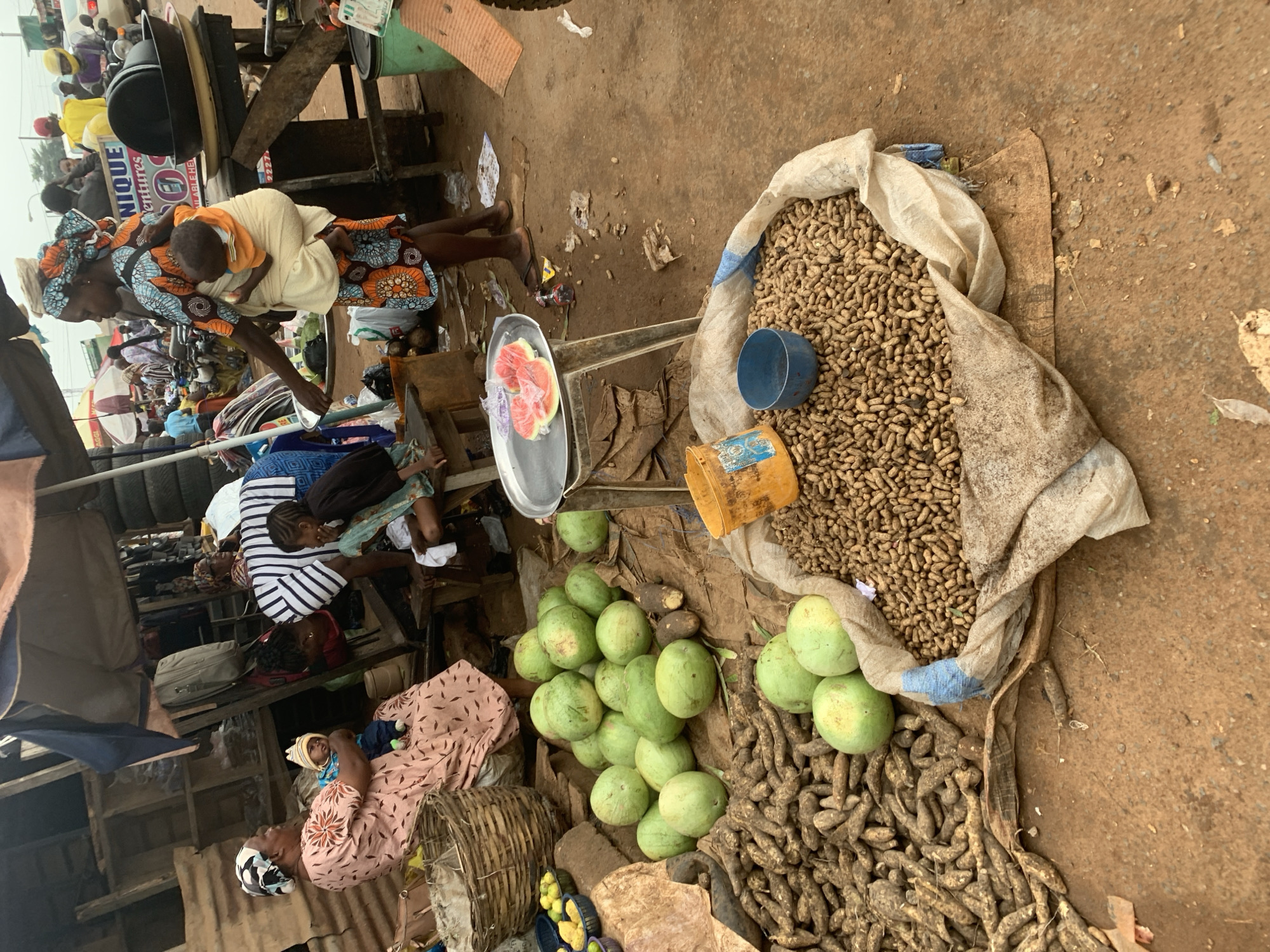 The image depicts a busy outdoor market scene in Shagamu, Nigeria. In the foreground, a large sack of peanuts rests on the ground, with a blue bowl placed on top. To the left, a pile of yams is visible, and further back, several round, green fruits, possibly watermelons or gourds, are scattered. In the mid-ground, a woman wearing a patterned dress and headwrap is carrying a baby on her back. She is standing near a stall with various goods. To her left, another woman in a pink dress and headscarf is seated, holding a baby. Behind them, a stall is set up with various items, including what appears to be tires and fabric. The ground is dusty and uneven, characteristic of an outdoor market. In the background, other market-goers and stalls are visible, along with a glimpse of vehicles, suggesting a bustling urban environment. The overall impression is one of daily commerce and community life in a Nigerian city.