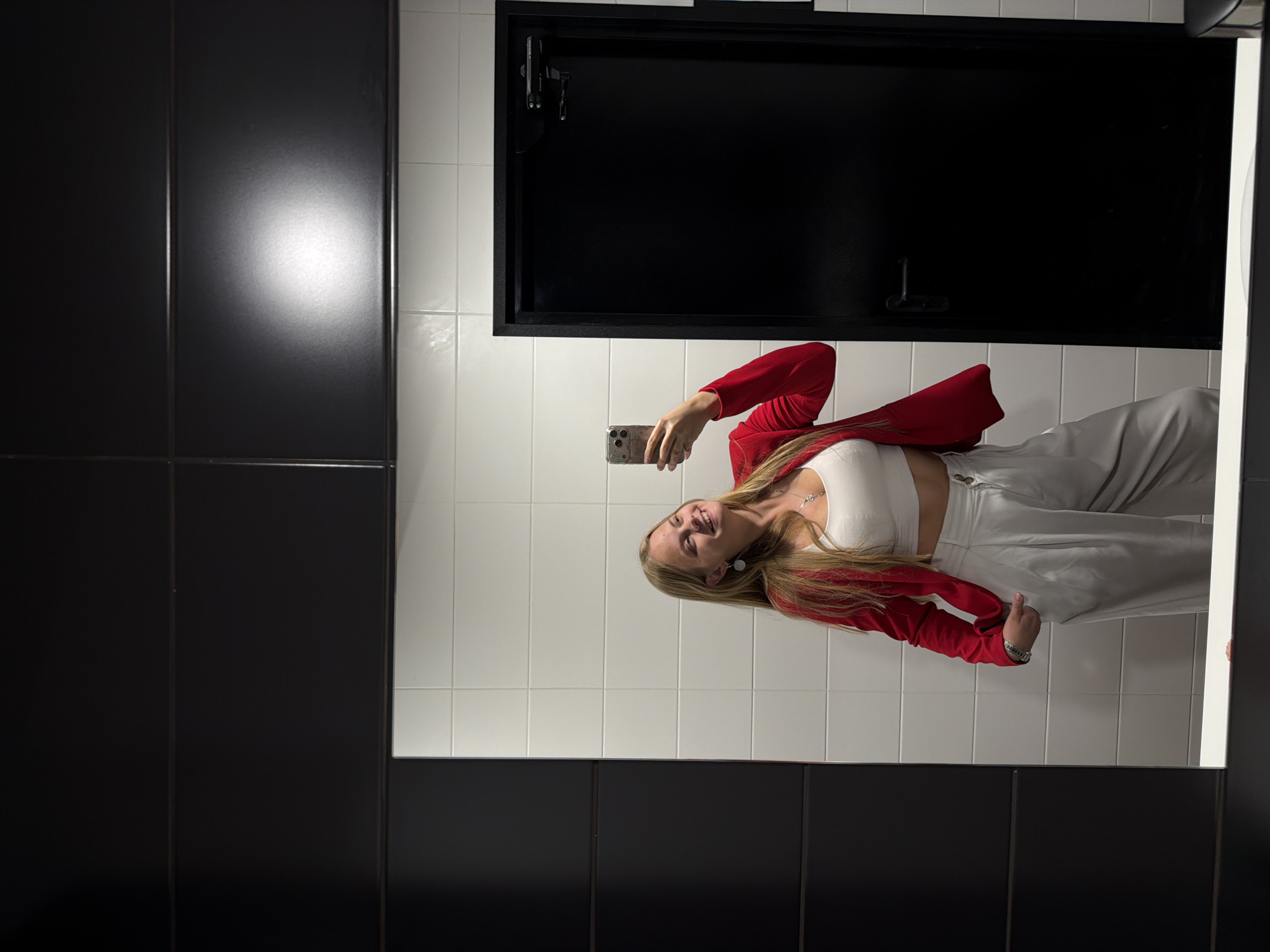 A young woman is taking a mirror selfie in what appears to be a modern restroom. She is wearing a white crop top, a vibrant red blazer, and light gray wide-leg pants. She is accessorized with a silver necklace and earrings. Her blonde hair is long and flows over her shoulders. The restroom has white tiled walls and a black door with a silver handle and a door closer. The overall aesthetic is clean and contemporary. The location is identified as Izhevsk, Russia.