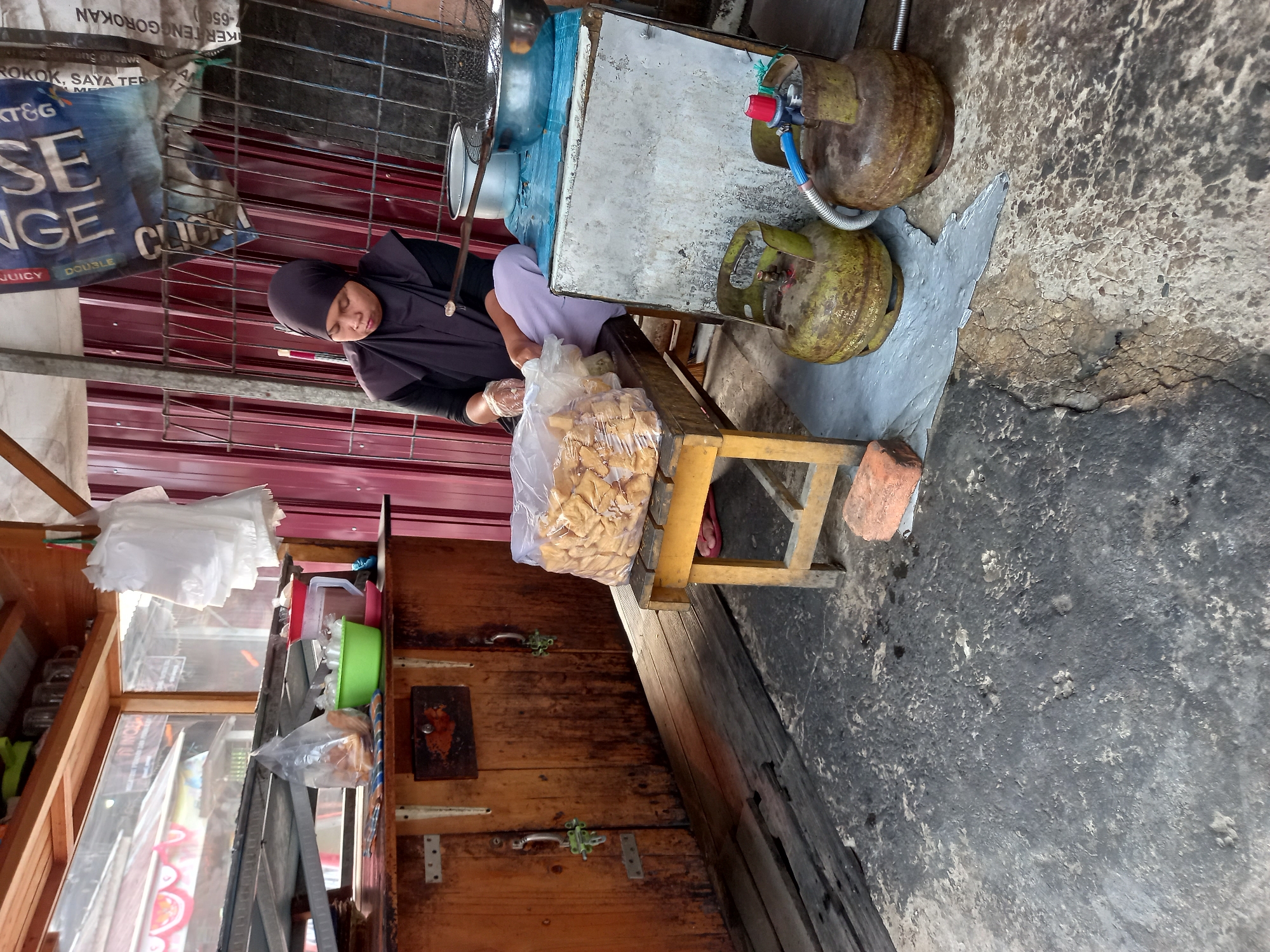 In the city of Samarinda, Indonesia, a woman wearing a dark hijab and light purple trousers is seated on a wooden stool at a street-side food stall. She is wearing clear plastic gloves and appears to be preparing food. In front of her, on the stool, is a large plastic bag filled with fried snacks, likely kerupuk, and a green bowl. She is positioned in front of a wooden counter, which has metal handles and a lock. Behind her, a corrugated red metal wall serves as a backdrop. To her right, on a blue surface, are two stainless steel bowls and a frying net. Two yellow gas cylinders are on the ground to the right of the stall, connected by a blue hose. The ground in front of the stall is rough concrete, with a weathered wooden plank pathway. A single red brick sits on the ground near the gas cylinders. The overall environment suggests a humble, outdoor food preparation and sales operation.