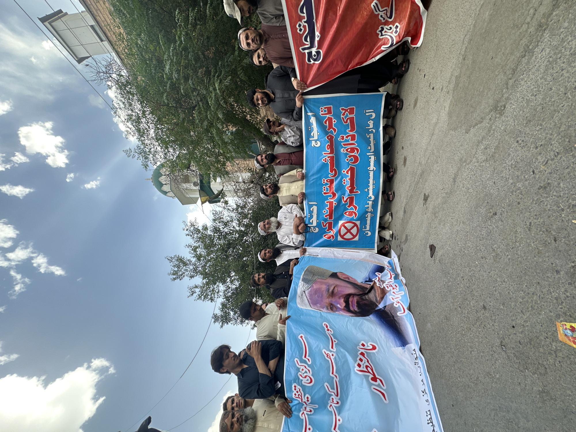 A group of men are standing together on a street, holding banners with Arabic text. The banners appear to be protesting something. Some of the banners also include a portrait of a man. The background shows buildings, trees, and a partly cloudy sky. Based on the text on the banners, the location is in Quetta City Tehsil, Pakistan.